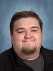 A high school teacher at FlexTech with glasses, a beard, and short dark hair smiling, wearing a green dress shirt and tie against a dark blue background.