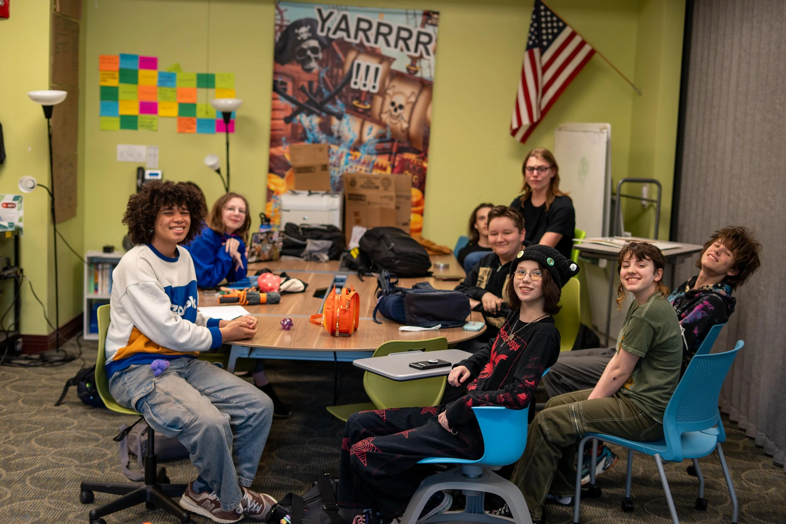 A group of young high school students sitting around a table in a classroom or meeting room, smiling and posing for the photo. 