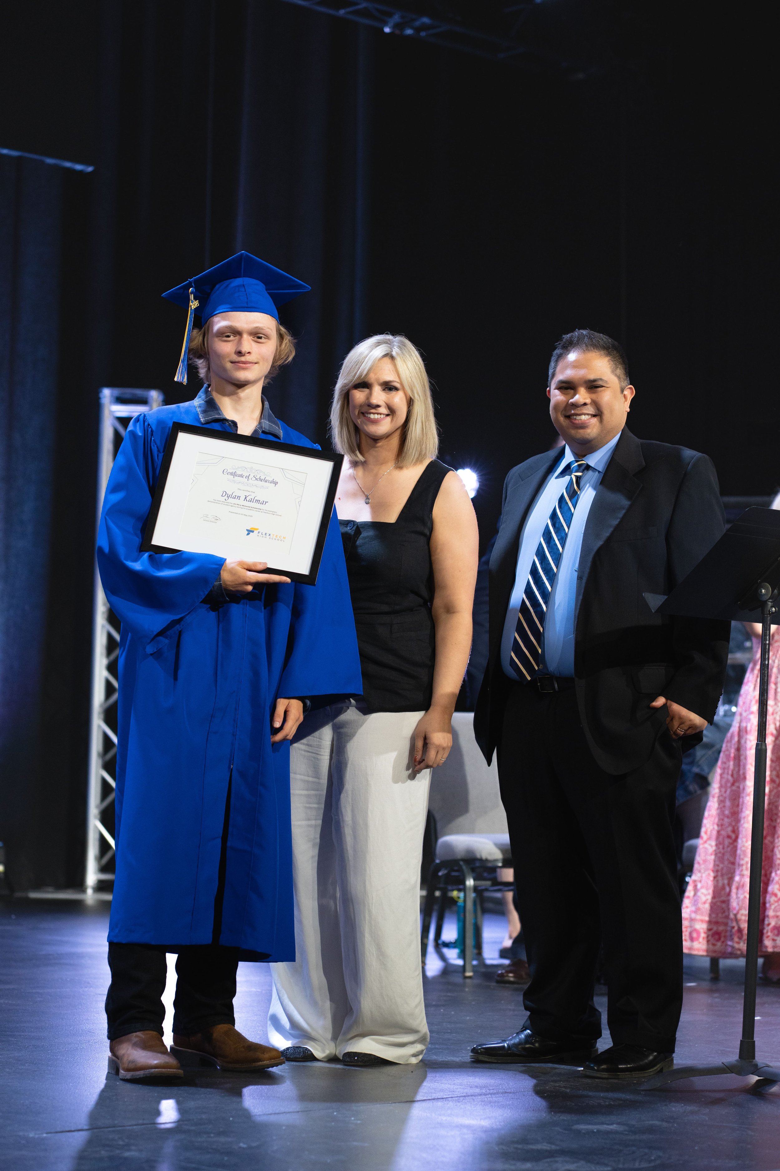 A young man in a blue graduation gown and cap holding a diploma, standing next to mom and dad on stage at FlexTech Brighton's graduation ceremony.