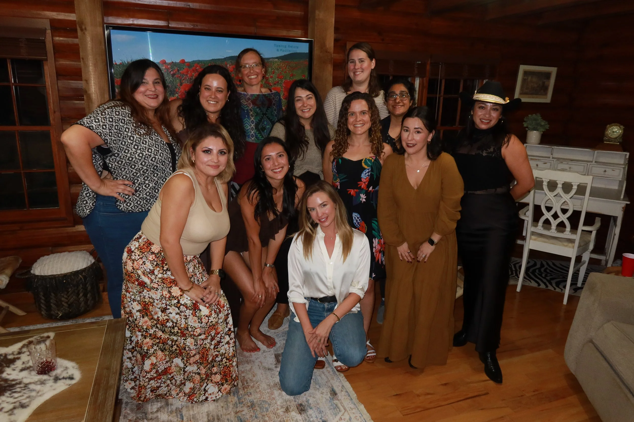 Group of 14 women smiling and posing together inside a wooden room with a large TV and furniture.
