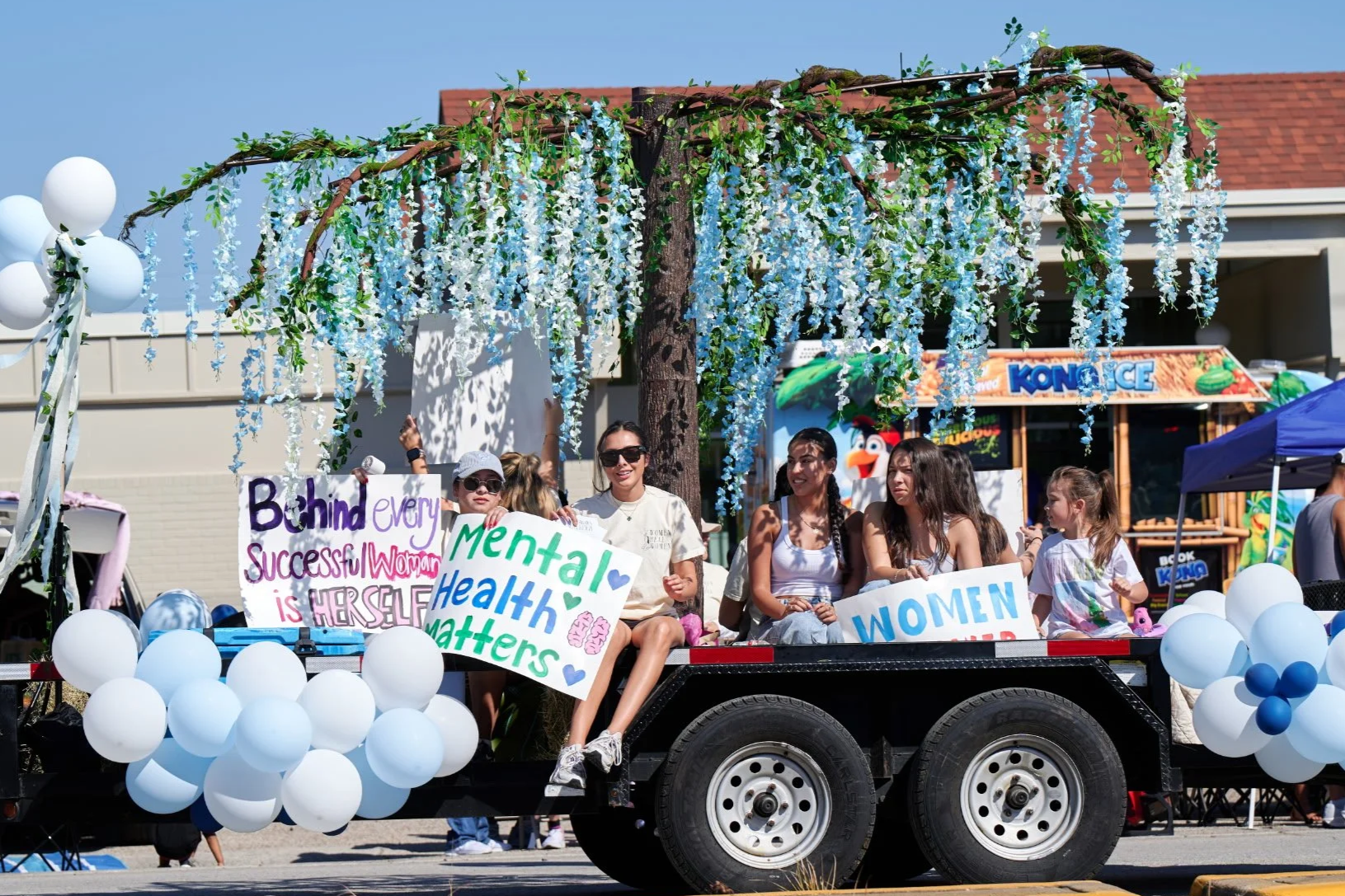 Women and girls sitting on a decorated float in a parade, holding signs supporting women’s mental health and success, with balloons and a large potted tree overhead.