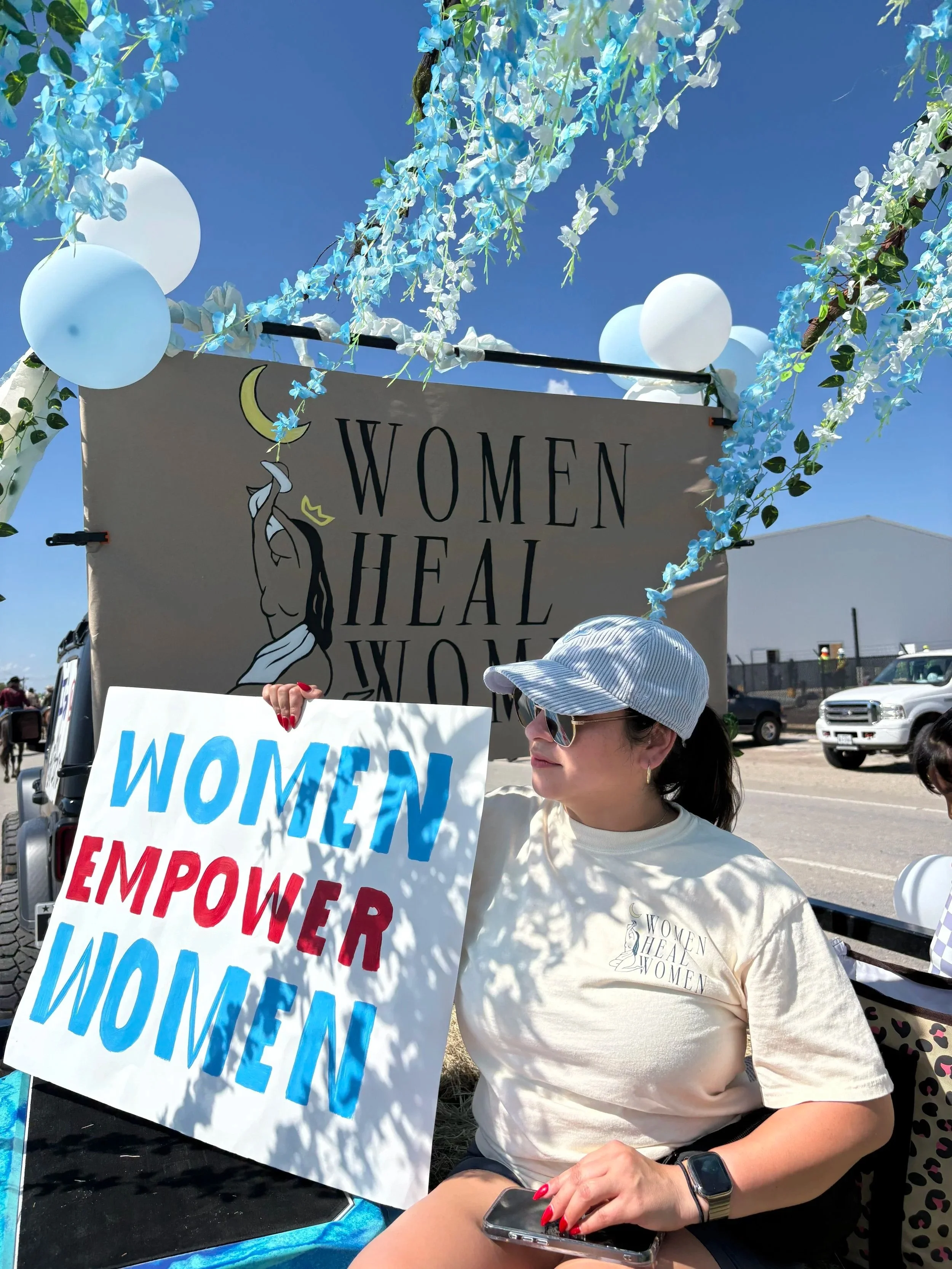 Woman holding a sign that reads 'Women Empower Women' at a women’s empowerment event. She is wearing sunglasses, a hat, and a T-shirt with the event's logo. Decorations include blue and white balloons and floral garlands.