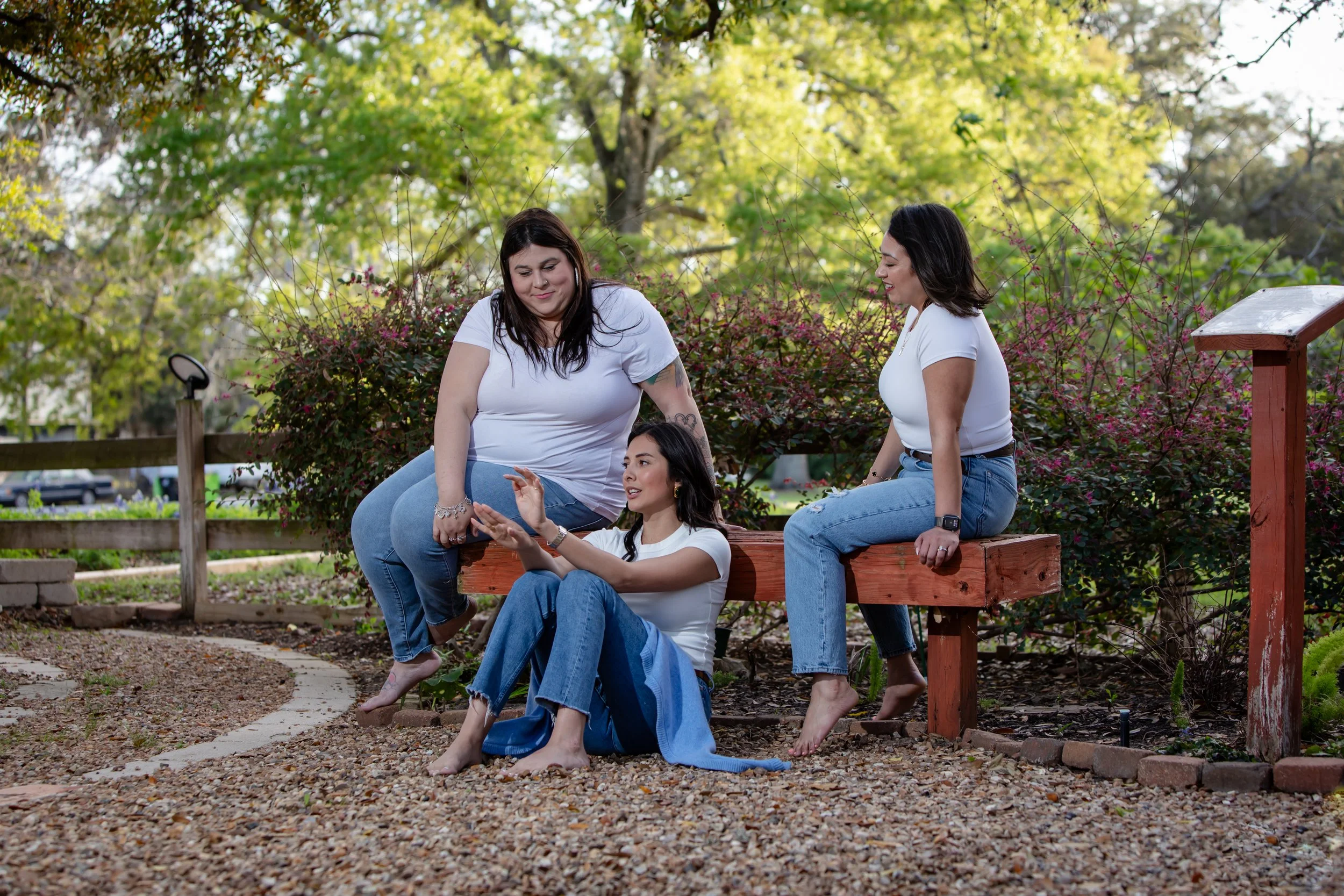 Three women sitting and chatting on a park bench surrounded by trees and plants.