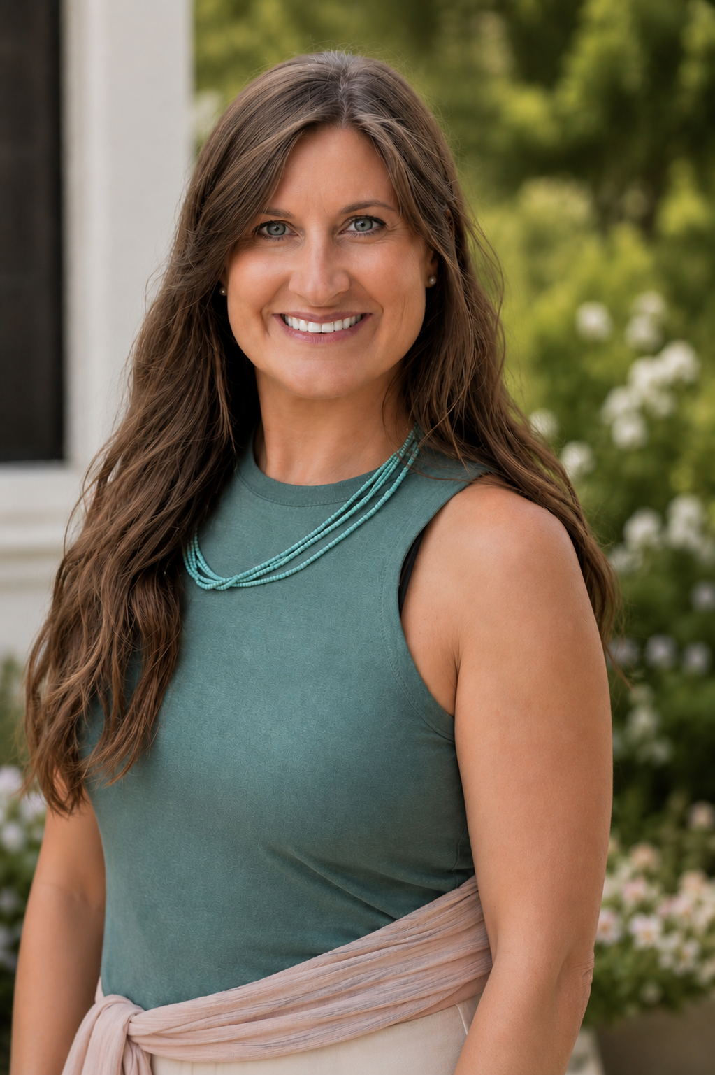 A smiling woman with long brown hair, wearing a teal sleeveless top and a layered turquoise necklace, standing outdoors with green trees and white flowers in the background.