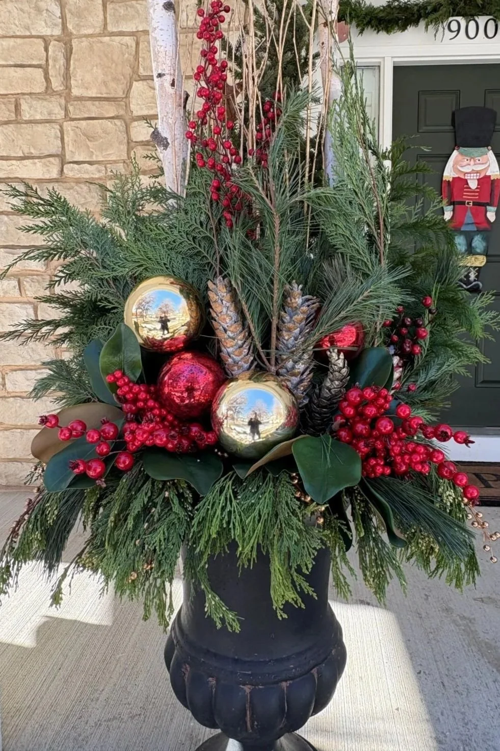 Holiday floral arrangement in a black vase with greenery, pinecones, red berries, and gold and red ornaments, set outside on a concrete surface with a brick wall and holiday decorations in the background.