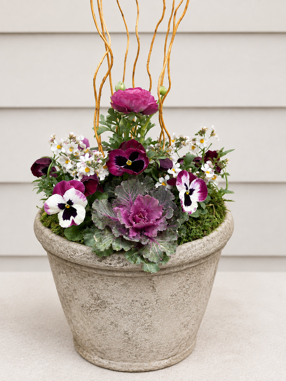 A decorative flower arrangement in a large textured beige planter contains purple and white pansies, pink cabbages, and other small white and purple flowers, with a light-colored background.