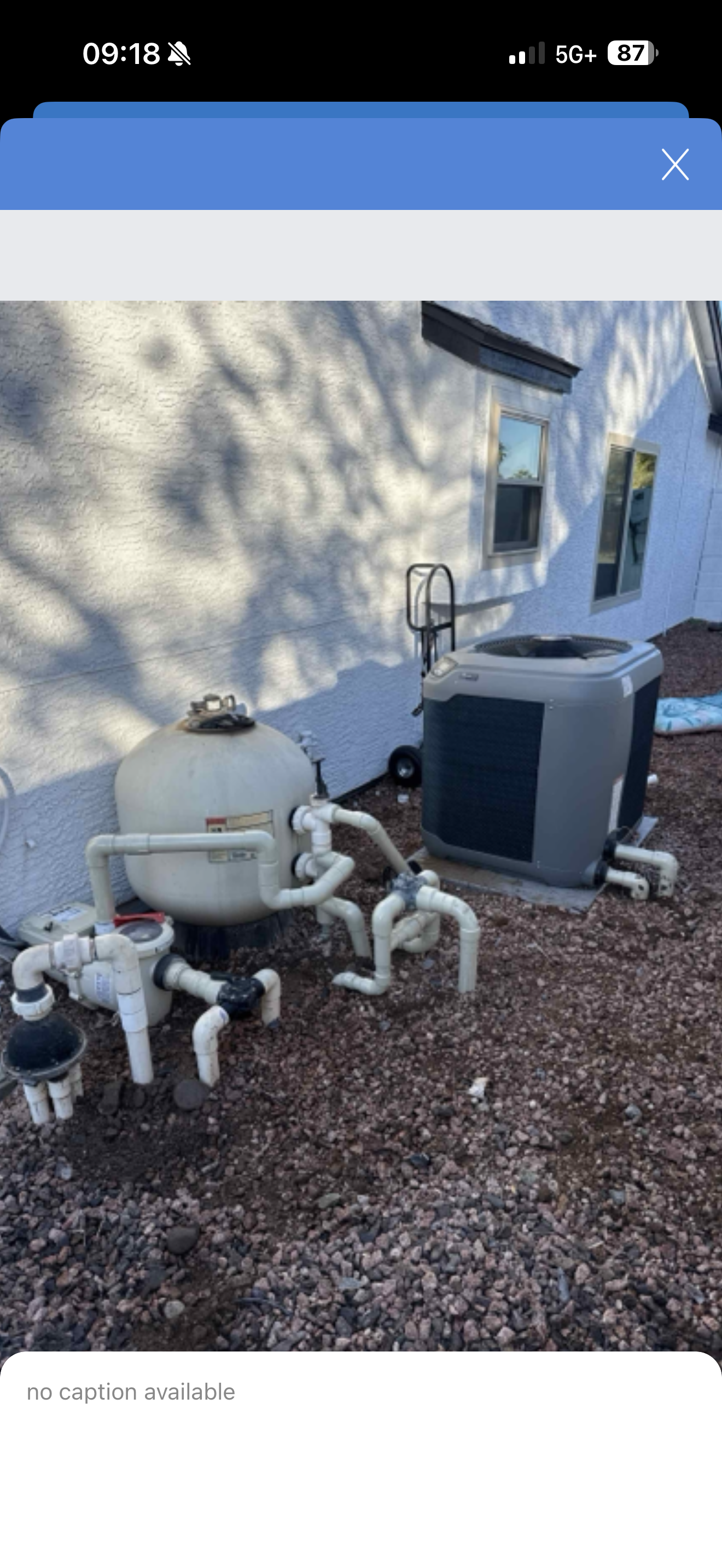 A backyard with a pool pump, filter, and filter valve connected with white PVC pipes against a white stucco house wall. There is gravel on the ground and shadow of a tree on the wall.