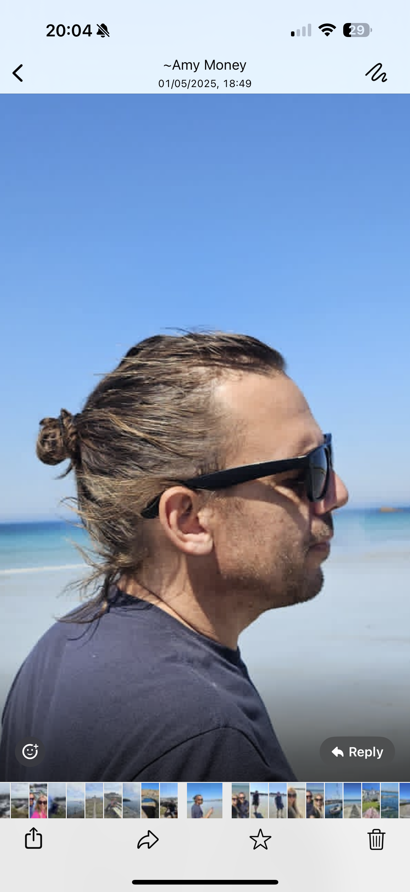 Profile photo of a man with long hair tied in a bun, wearing sunglasses, looking to the right at a beach with blue sky and ocean in the background.