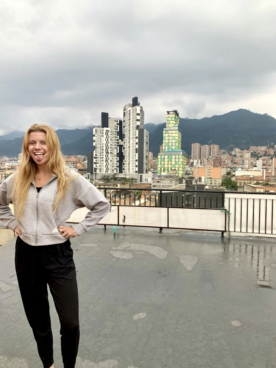 A young woman with long blonde hair, wearing a gray zip-up hoodie and black pants, standing on a rooftop with a city skyline and mountains in the background on a cloudy day. She is smiling and sticking her tongue out.
