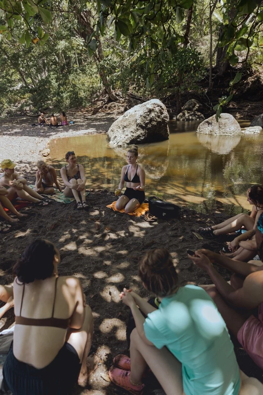 A group of people gathered on a dirt patch near a river, listening to a woman in a black swimsuit who is sitting on an orange towel and speaking to them. The scene is shaded by tree branches, with large rocks and water in the background.
