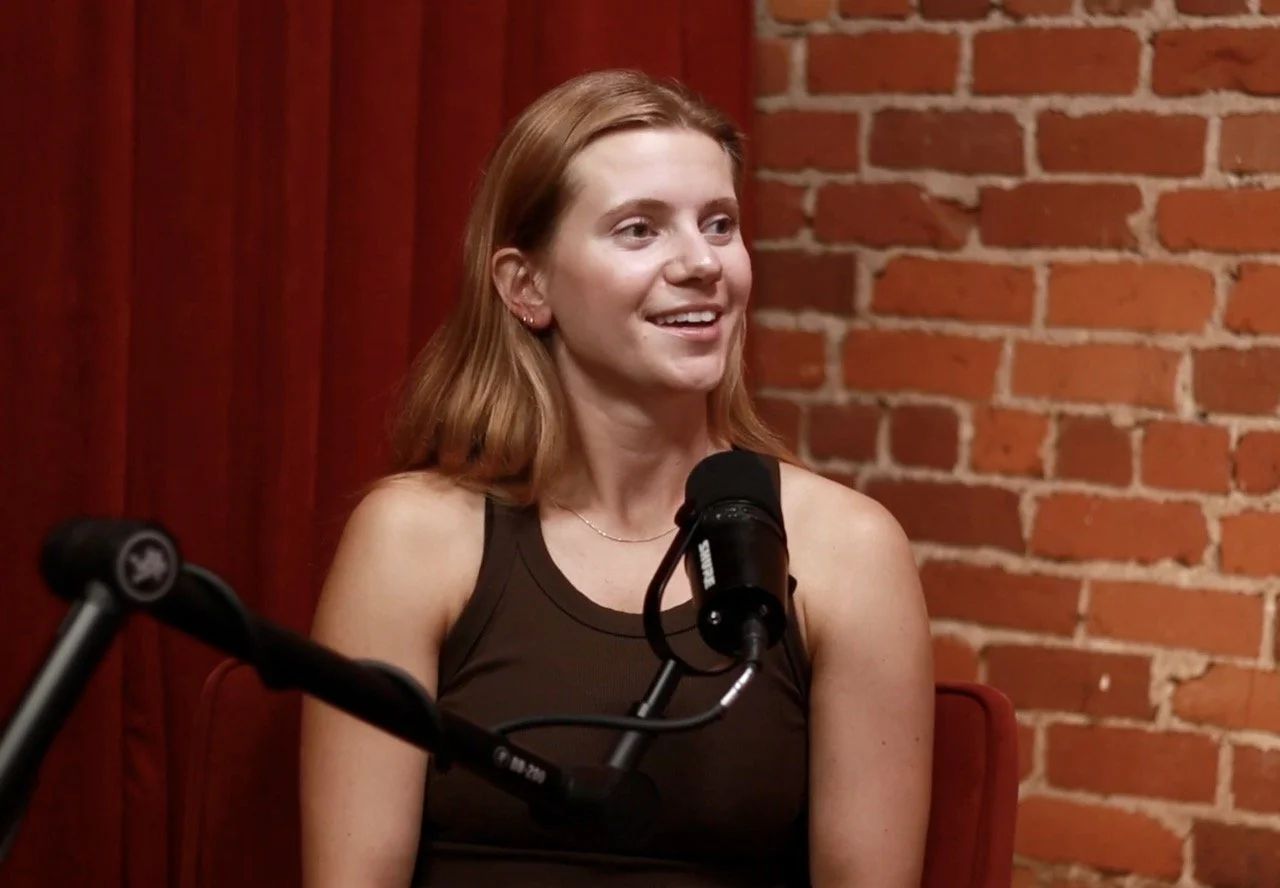 A woman with shoulder-length red hair, wearing a sleeveless black top, sitting in front of a microphone, with a brick wall and red curtain in the background, speaking or listening during a recording session.