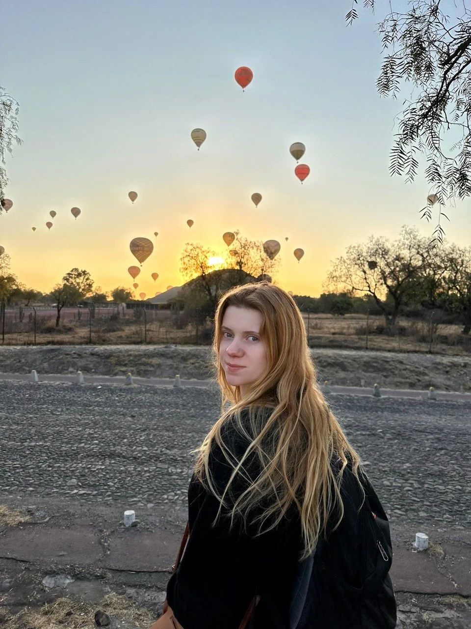 A young woman with long blonde hair standing outdoors at sunset with hot air balloons in the sky behind her.