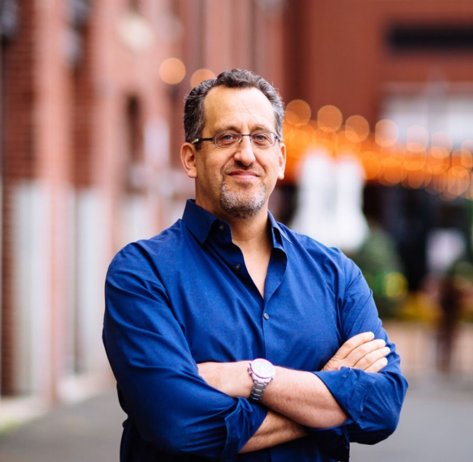 Man wearing a blue shirt with crossed arms standing outdoors with a blurred background.