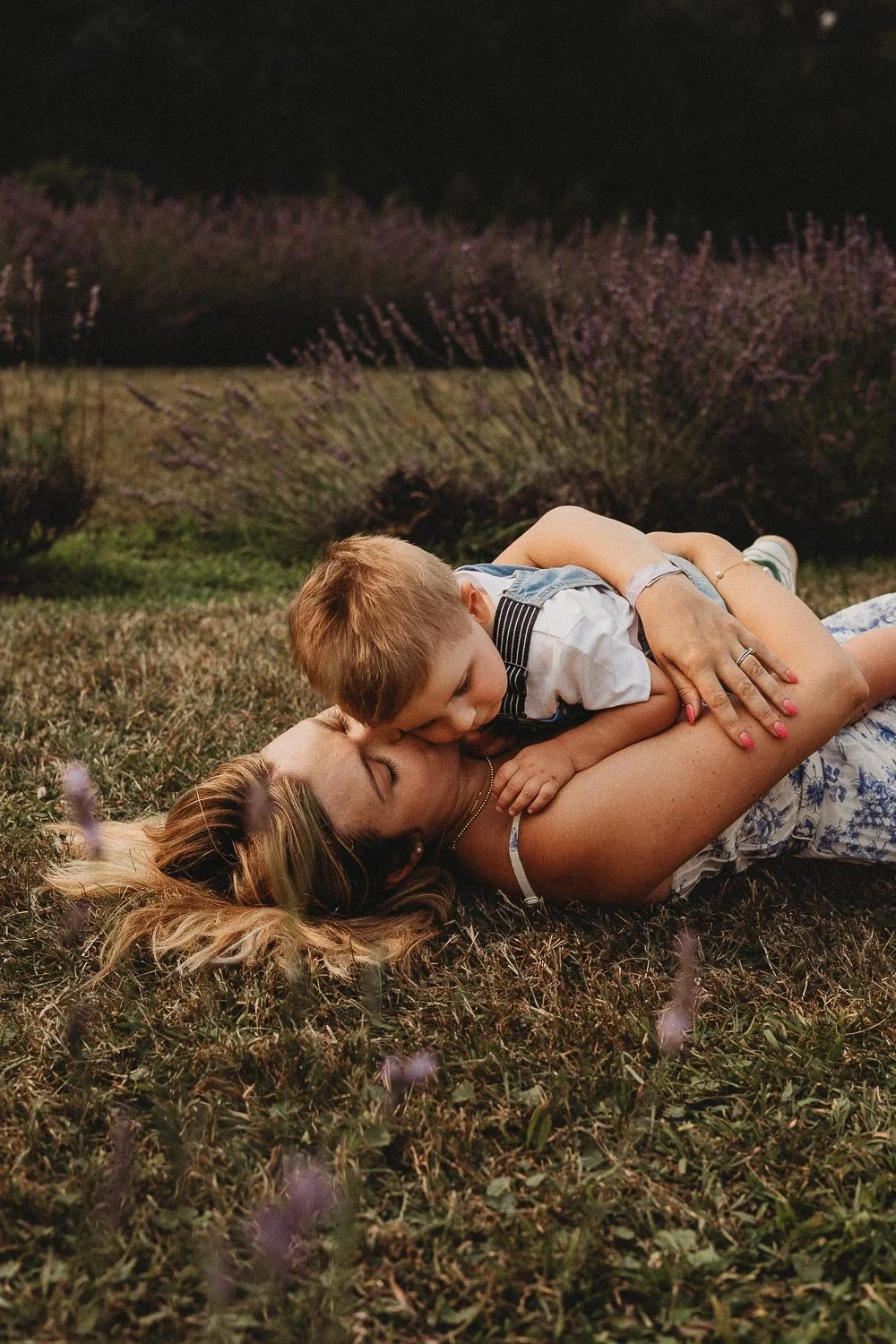 A woman and young child lying on the grass outdoors at night, hugging and kissing during a family photoshoot at La Maison Lavande, Quebec