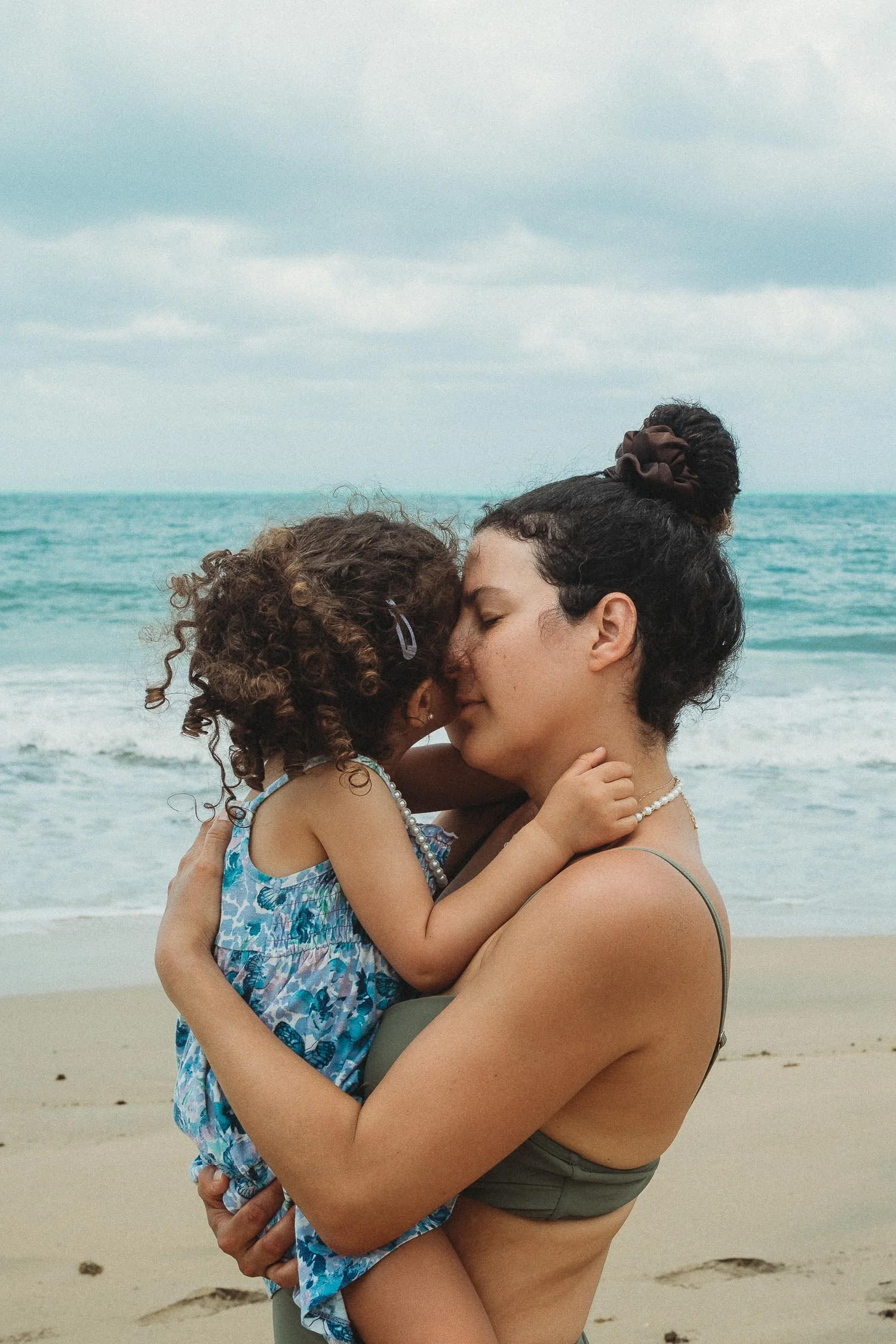 Jen Blanco and daughter embracing on a beach, with their foreheads touching, while the ocean and cloudy sky are in the background.
