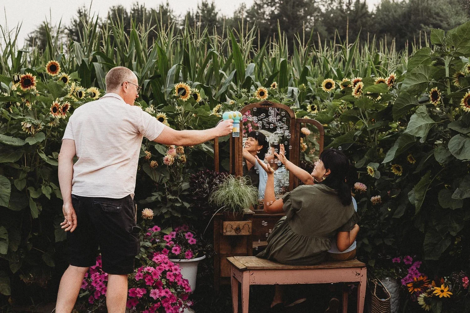 A man is blowing bubbles towards two women sitting in front of a mirror, surrounded by sunflowers and pink flowers in a garden during an outdoor family photo session.