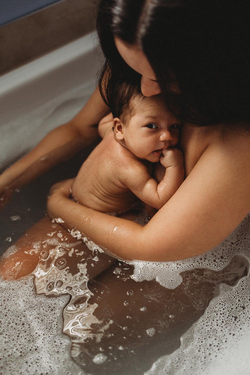A woman holds a baby in a bathtub, with the baby resting on her chest and looking at the camera, while she gently supports the baby during bath time.