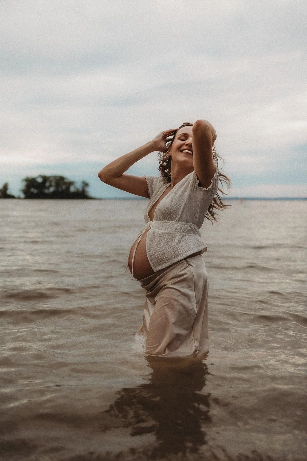 Pregnant woman flipping her hair and smiling while standing in the water at an outdoor maternity photoshoot in Cap-Saint-Jacques, Montreal