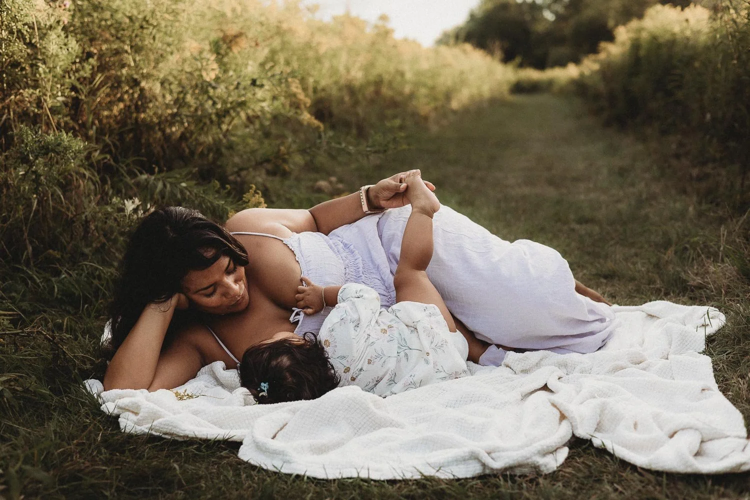 Mom laying down on a blanket with her toddler daughter, touching her little foot during an outdoor family photo session at Boucherville Island