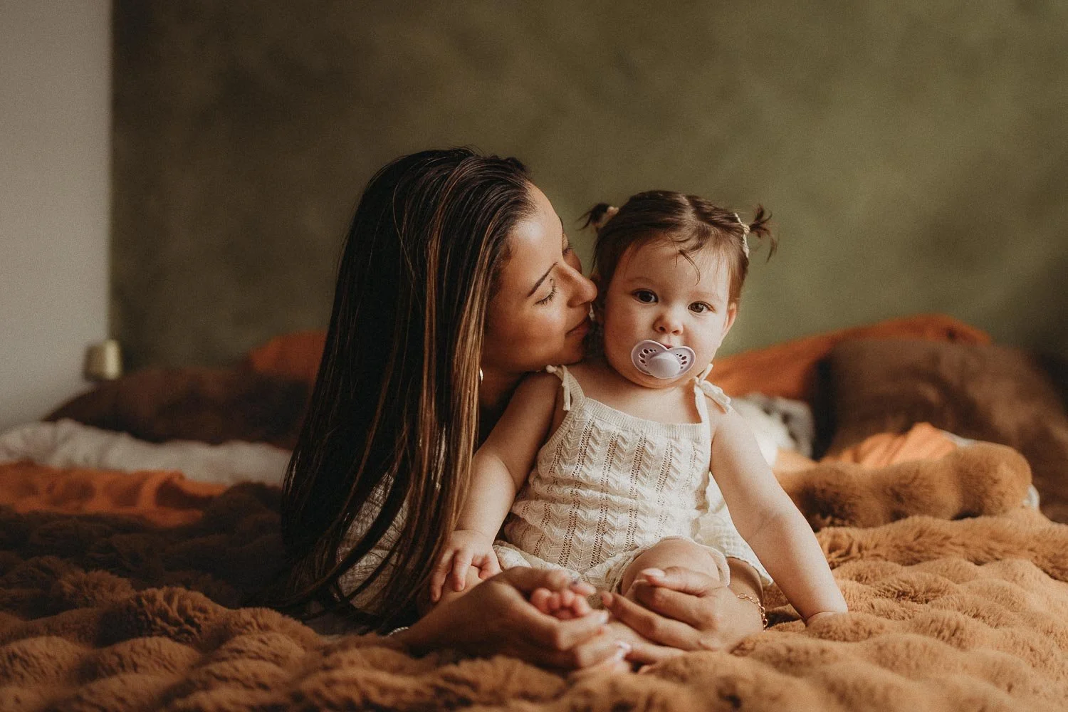 Mom laying down in bed next to her seated daughter. Mom is almost kissing her baby while the kid looks at camera. Captured at an in-home Motherhood Photoshoot in Montreal