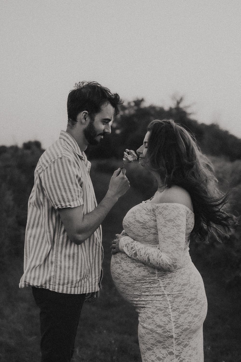 A black & white photo of a pregnant mom gently touching her belly smelling flowers that her male parter is offering, while both smiling, captured during an outdoor maternity photoshoot in Boucherville Island, Montreal