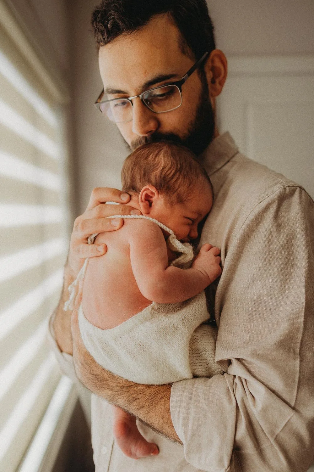 A man with glasses and a beard holding a newborn baby close to his chest, both with closed eyes, near a window with blinds.