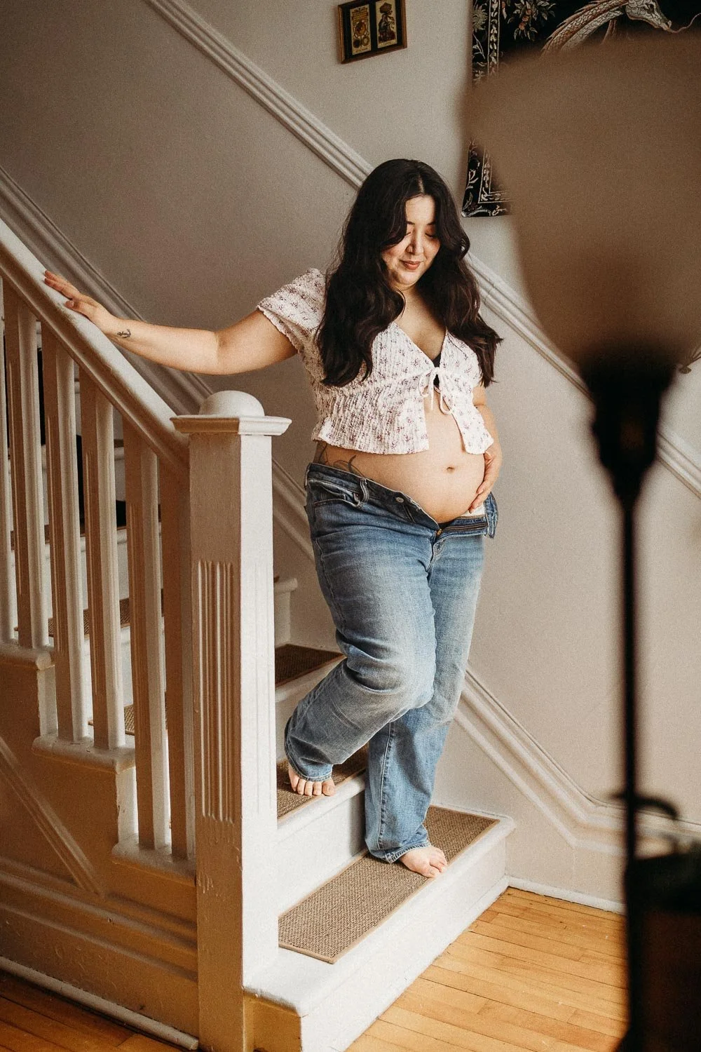 A pregnant woman with dark hair standing on stairs, holding her belly, wearing a cropped top and blue jeans, smiling and looking down during an in-home maternity photo session in Dorval, montreal