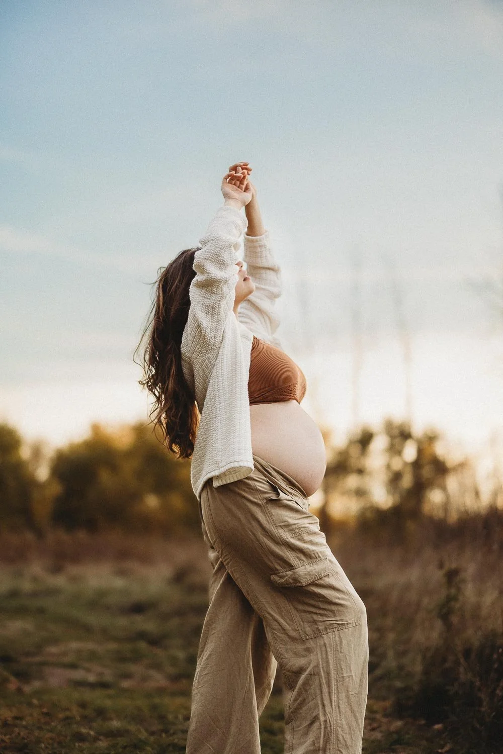 Pregnant woman outdoors at sunset, stretching her arms above her head in a field captured during an outdoor maternity photoshoot in Boucherville Islands near Montreal
