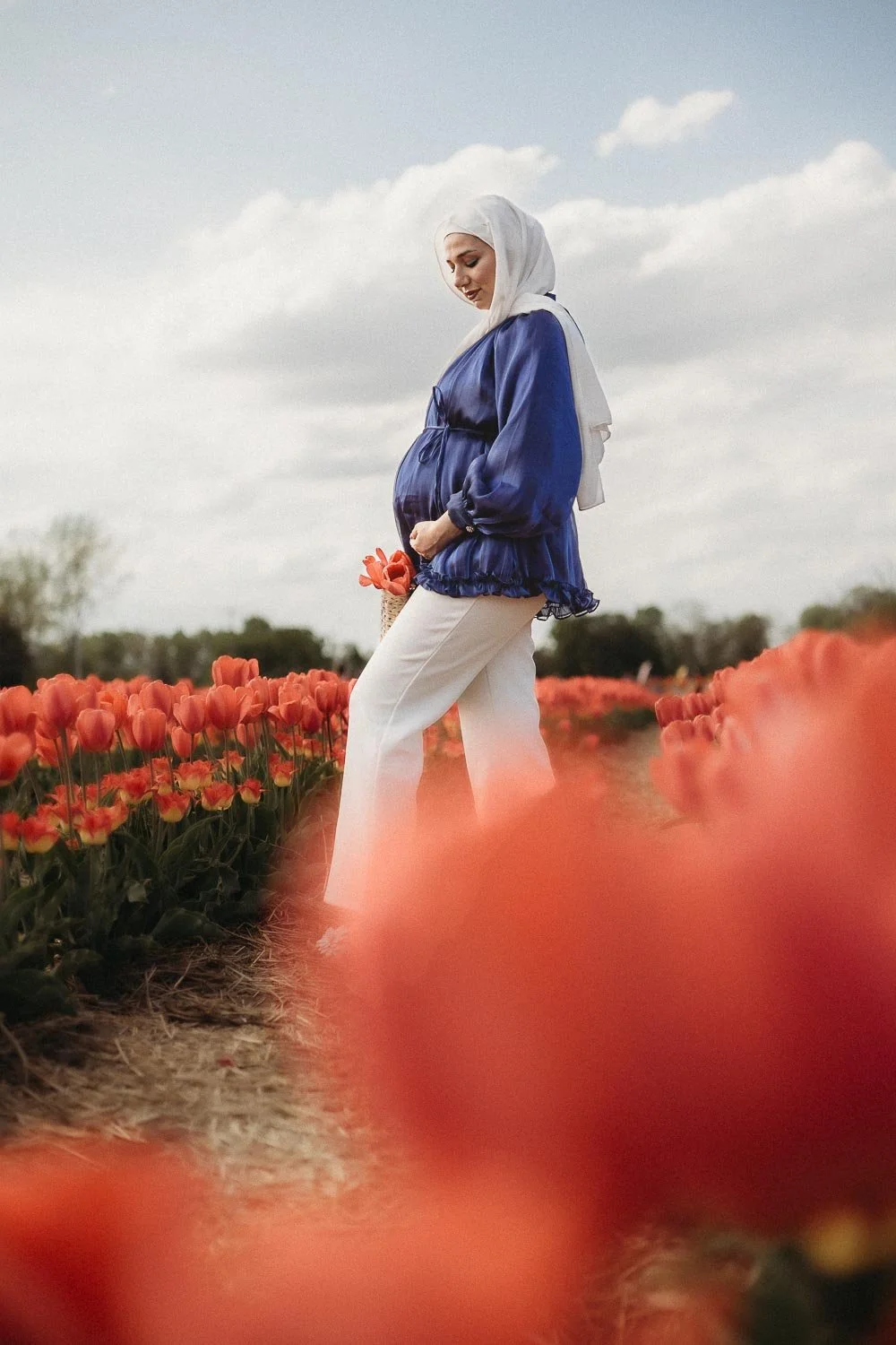 A pregnant woman with light skin wearing a white head scarf, a blue jacket, and white pants standing in a tulip field holding a small basket of flowers, with a cloudy sky background captured at a Maternity photoshoot in the Tulip Fields, Laval
