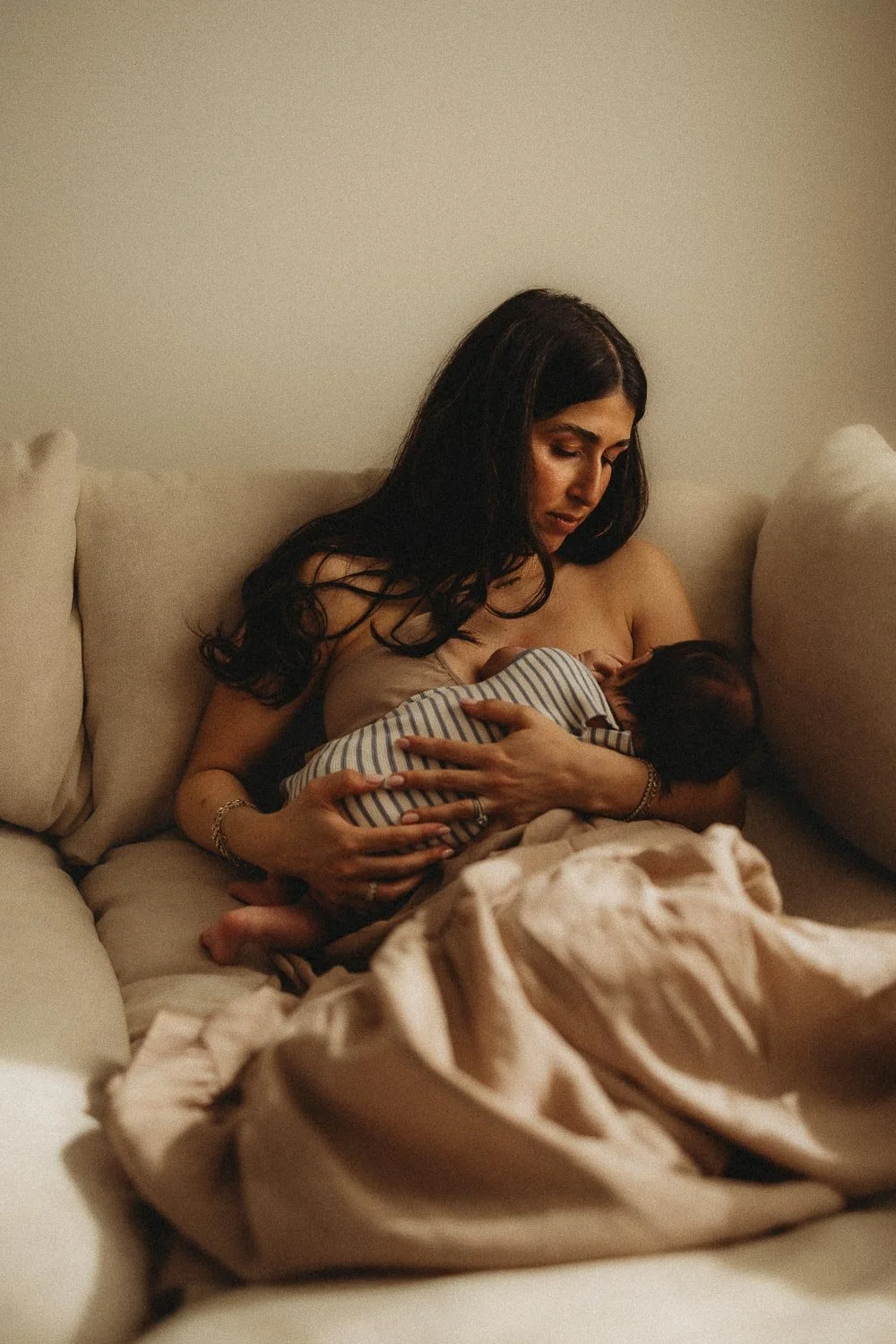  Mom breastfeeding her baby in the corner of a couch dressed in a soft beige dress, light illuminating the scene beautifully, captured at a Montreal in-home newborn photoshoot. 