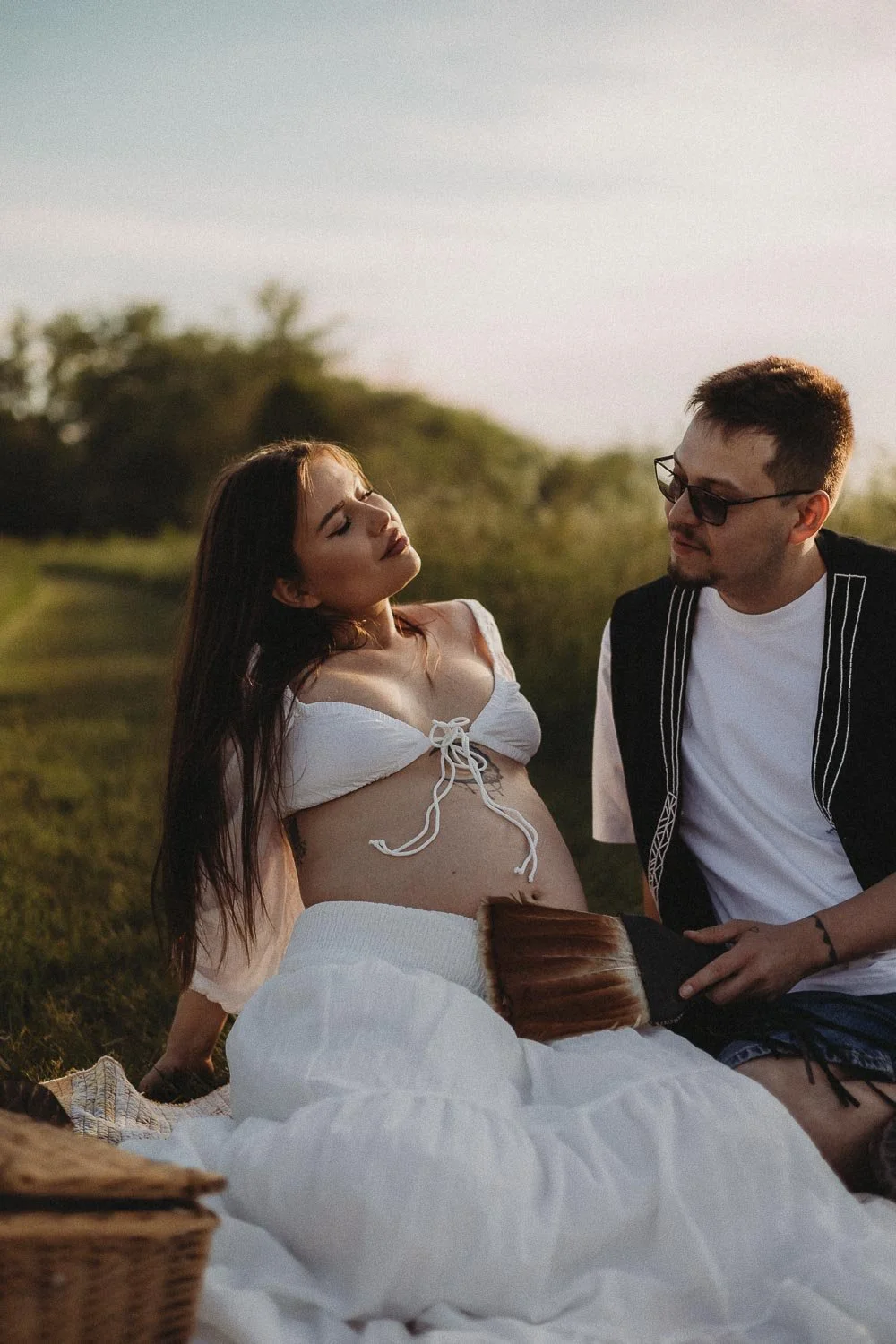 Pregnant woman and partner seated and looking at each other while the man is holding a Mohawk fan on her belly during an outdoor maternity photoshoot in Boucherville Island, Quebec