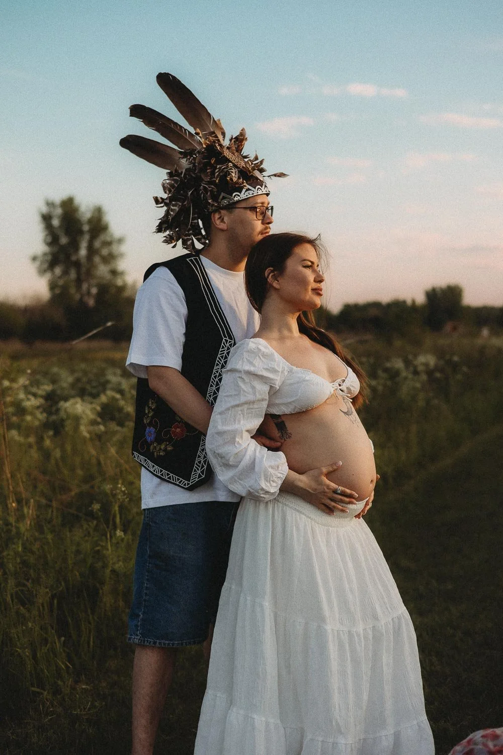A pregnant woman from the Mohawk community being held by her partner, both looking at the horizon captured at golden hour during an outdoor maternity photo session in Boucherville Island