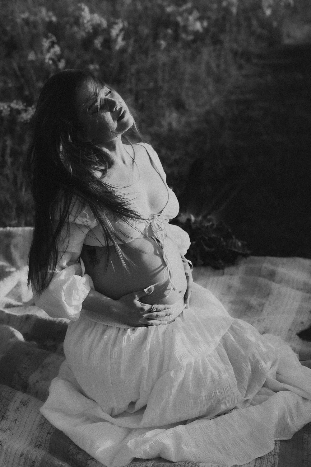 A black &amp; white photo of a pregnant woman captured in mid head movement with hair in her face and closed eyes while holding her belly at the Boucherville islands during a maternity photoshoot