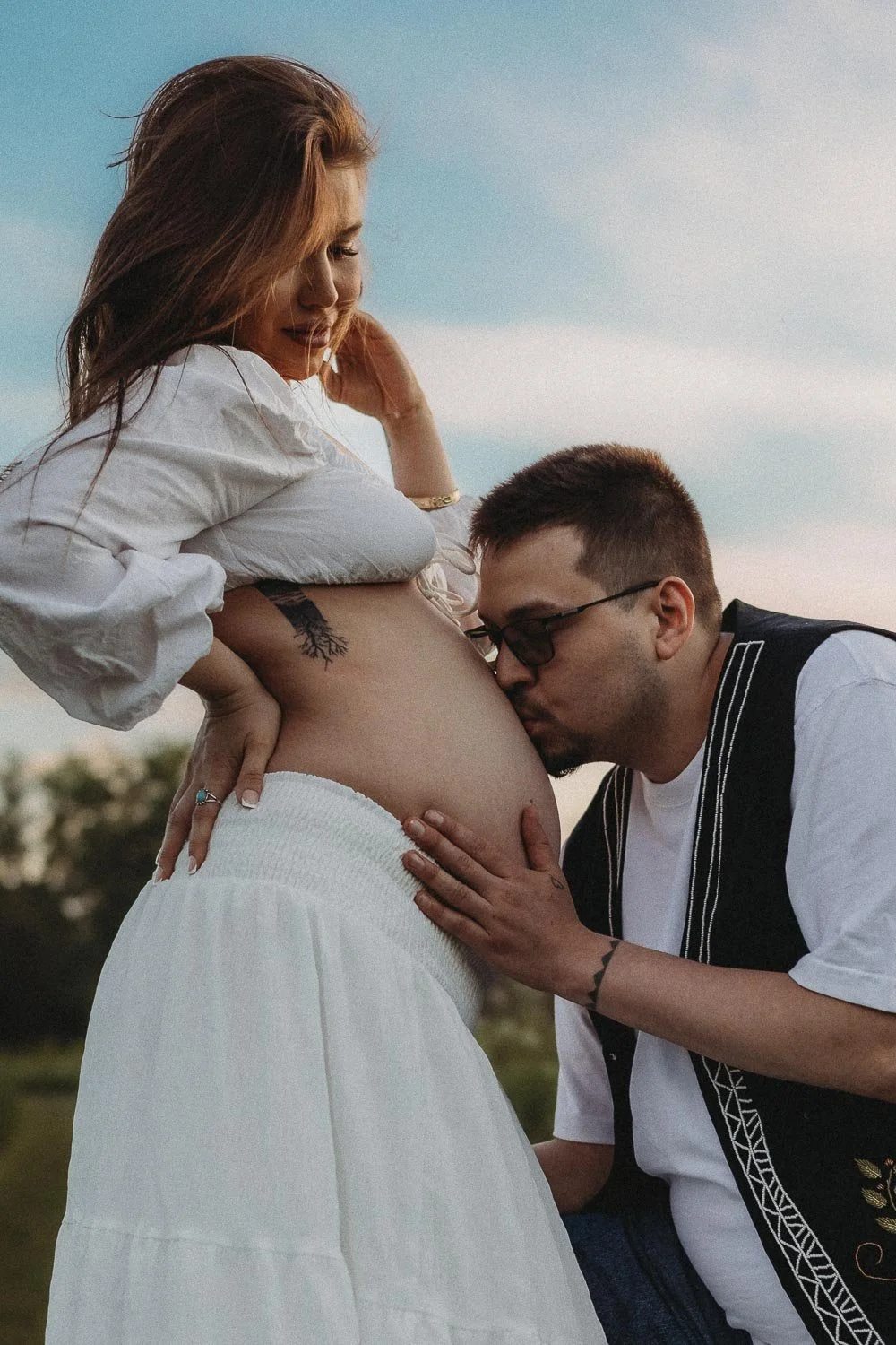 A pregnant woman looking at her partner who is kneeling and kissing her pregnant belly during an outdoor maternity photoshoot in Boucherville Island