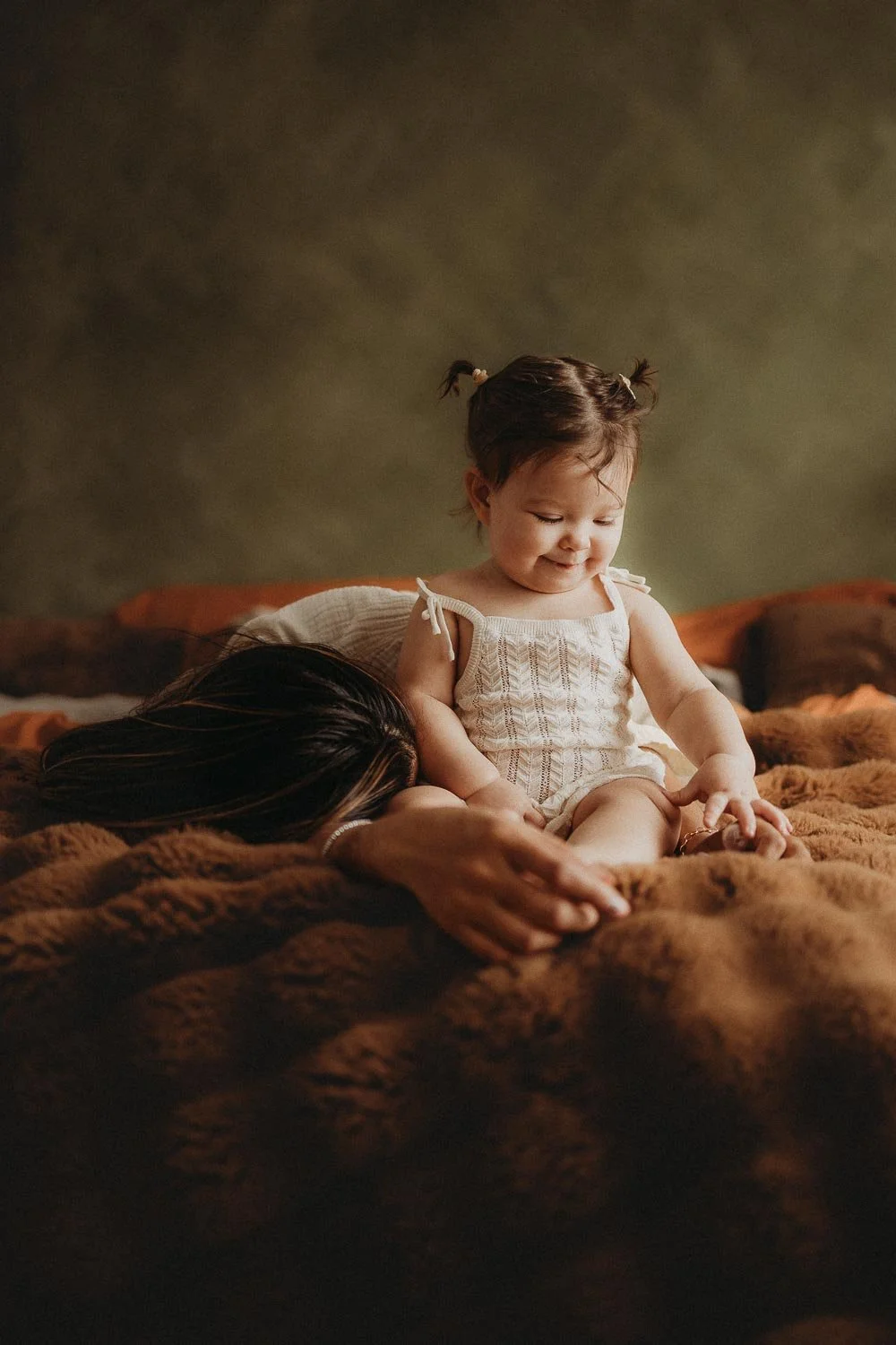 Mom laying down next to her baby girl while the little one is sitting, smiling and looking at her knee, captured during an in-home Motherhood Photo Session in Montreal