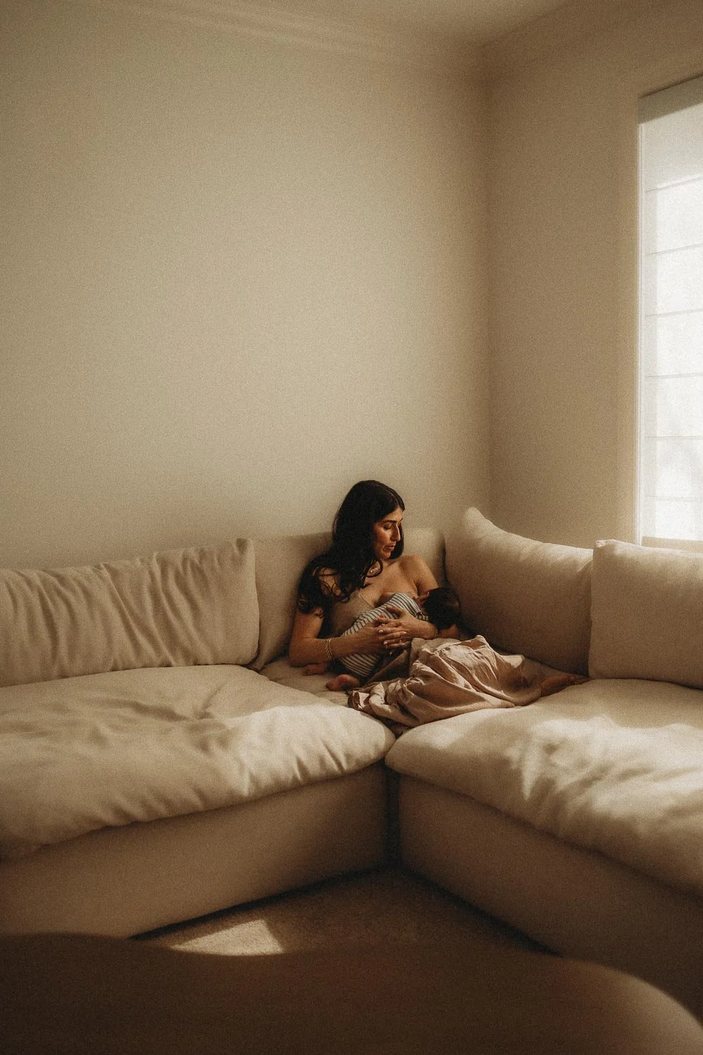 Mom breastfeeding her baby comfortably at the corner of her couch durin ga sunny day which let's in sun light creating nice contrast, captured during an in-home newborn photo session in Hampstead, Montreal