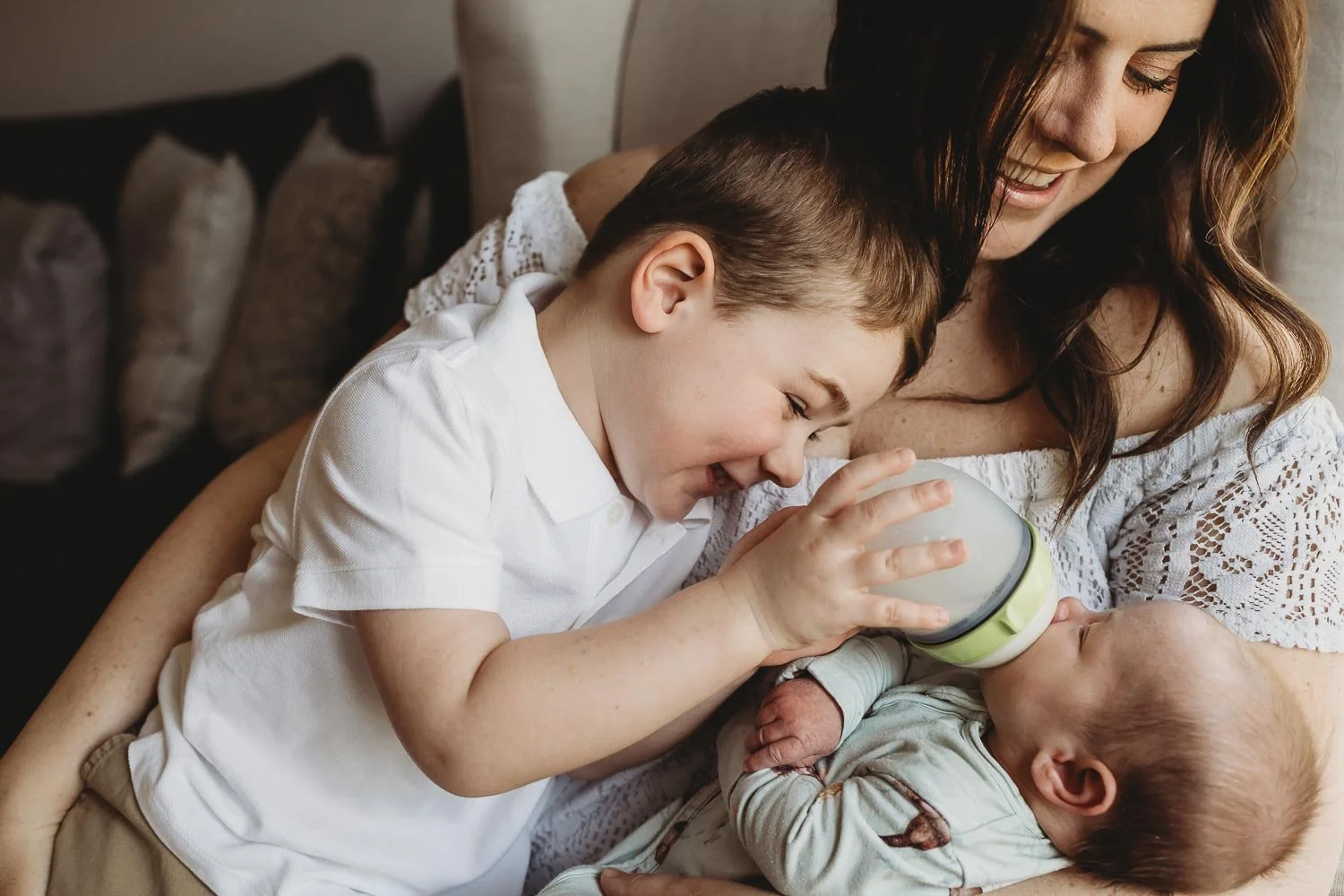 Mom with her 2 kids, a toddler and newborn. The toddler is feeding the baby a bottle and mom is smiling, captured during an in-home newborn photo session in TMR Montreal