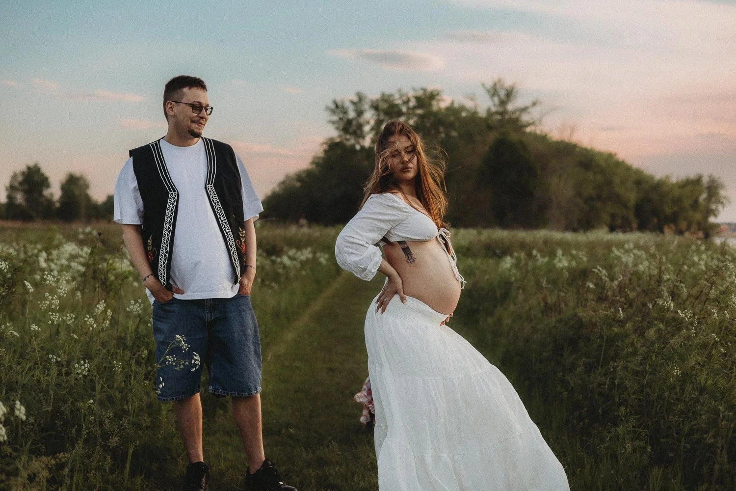 A pregnant woman holding her back in a sexy way while her husband is behind her looking at her with a smirk on his face, both in the middle of nature at a beautiful pink sunset, captured during an outdoor maternity photoshoot in Boucherville Island
