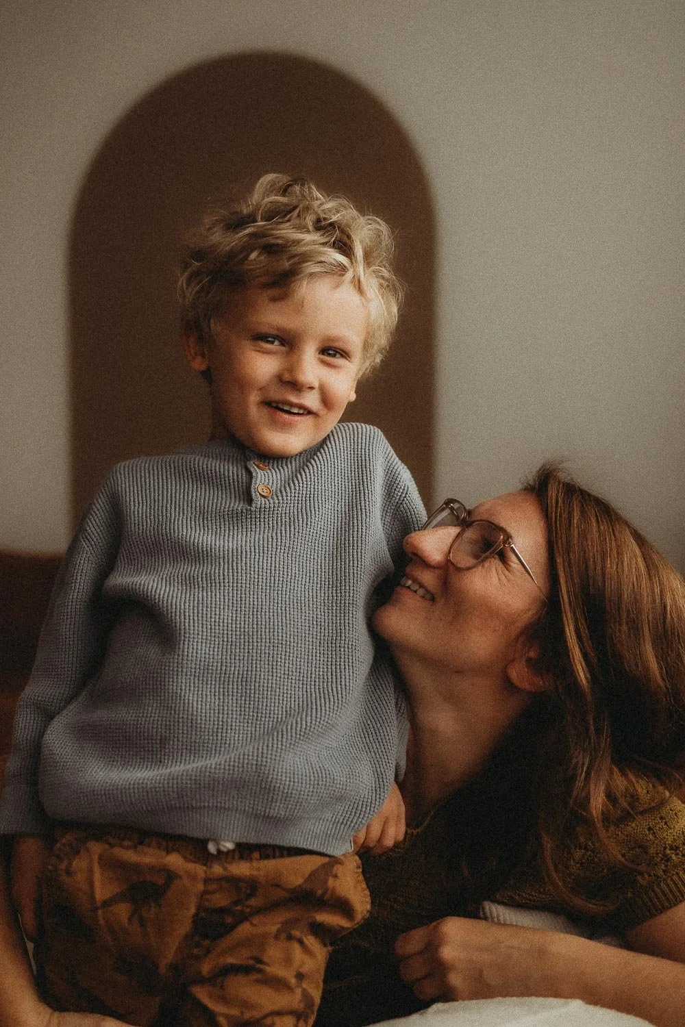 A smiling woman with glasses looking at a young boy with tousled blonde hair, who is standing and wearing a gray shirt. They are indoors, and the woman is lying on her stomach beneath the boy.