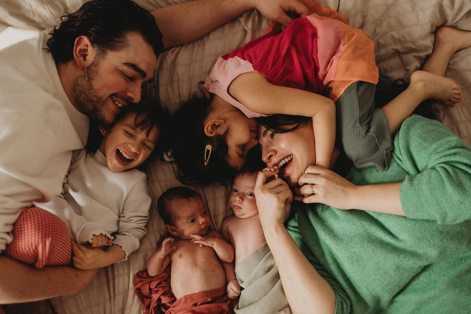 A family of 6 laying down in bed and laughing, mom, dad, 2 sisters and newborn twins captured at an in-home photoshoot in La Salle, Montreal