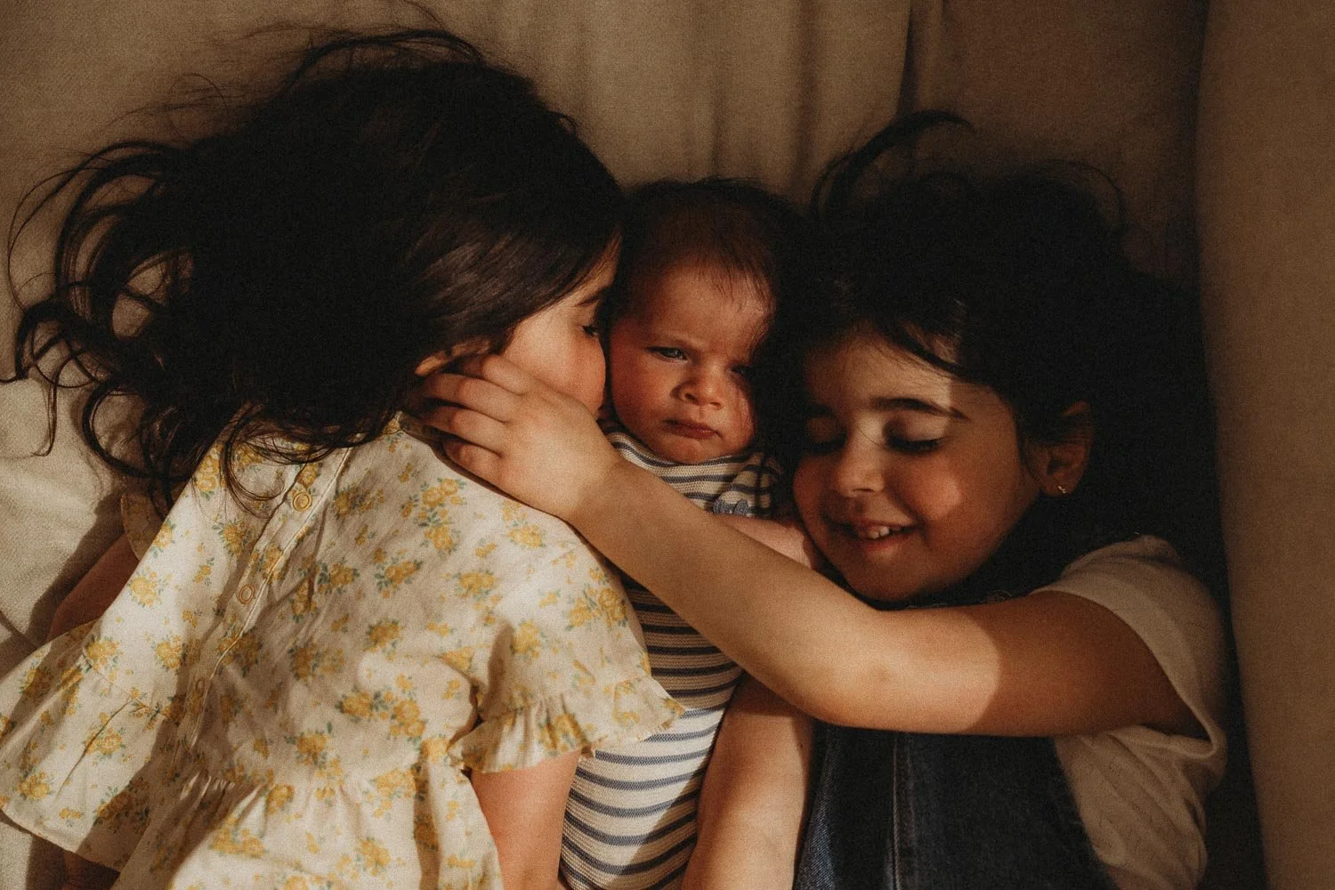 Two girls and their baby brother laying down and cuddling on a sofa while the sun light hit their faces beautifully, captured during an intimate Montreal in-home newborn photoshoot