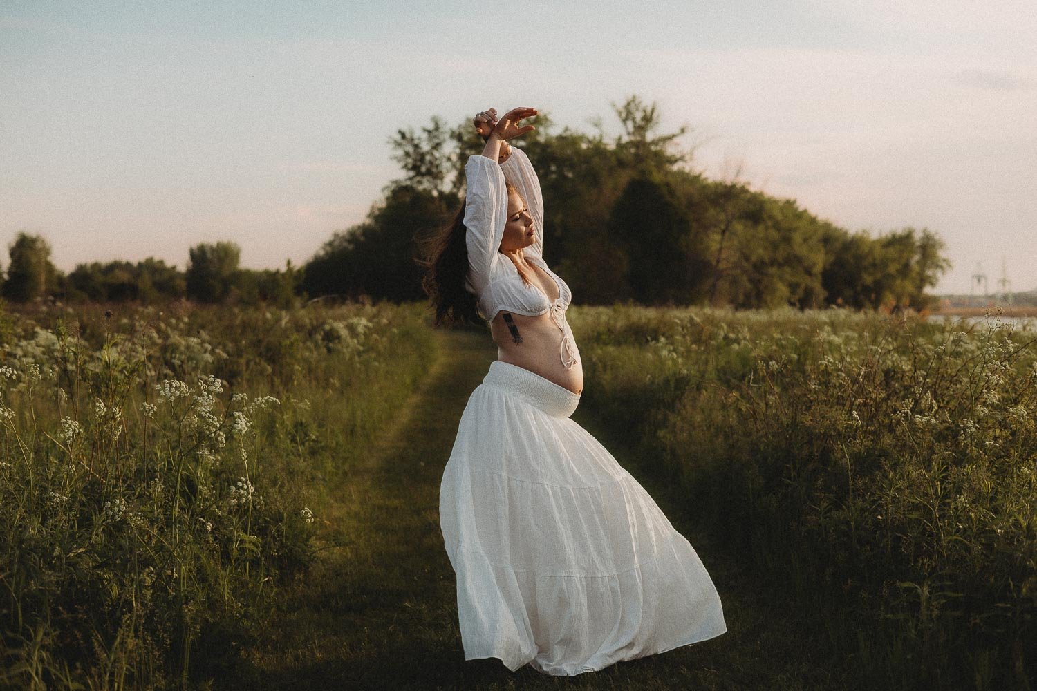 A pregnant woman lifting her arms and looking into the horizon in the middle of nature wearing a white skirt and crop top  captured during an outdoor maternity photoshoot in Boucherville Island, Montreal