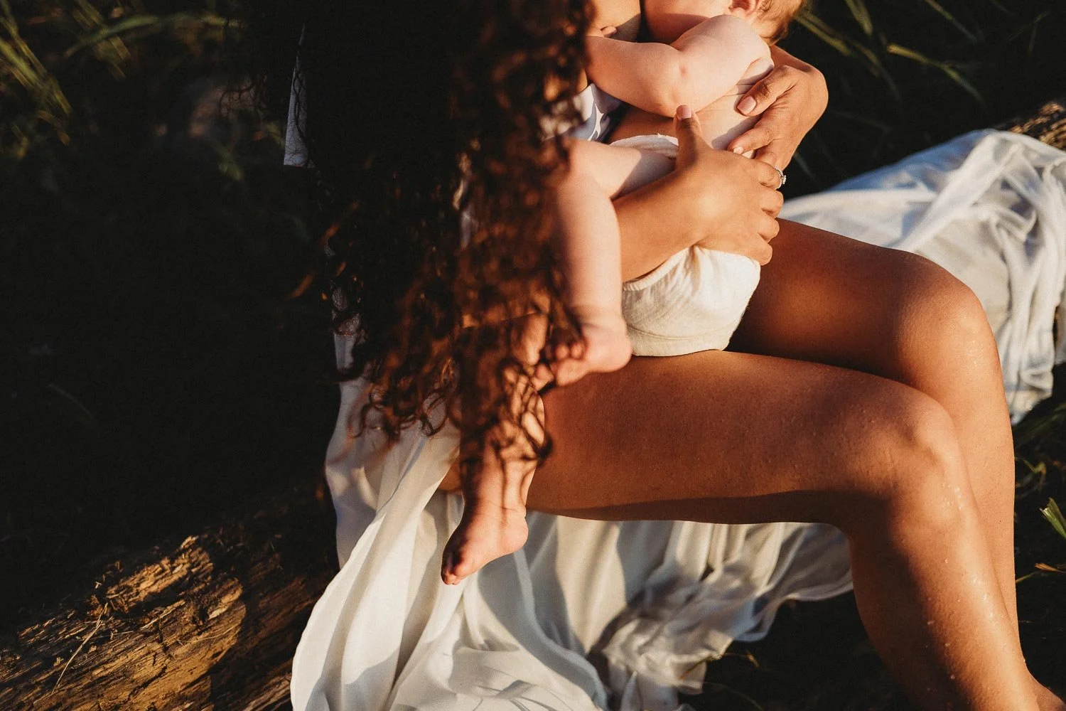 An outdoor motherhood photoshoot at Cap-Saint-Jacques portraying a mom holding her baby while breastfeeding her and sitting on a driftwood log outdoors, with a white cloth draped around them, during sunset.