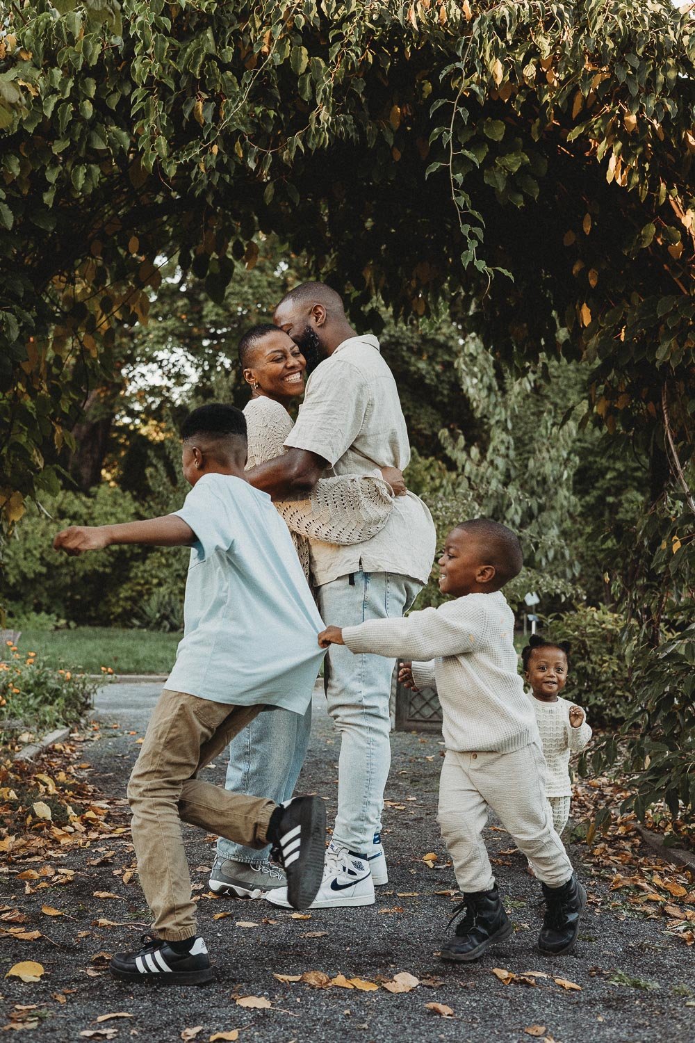A happy Black family of five enjoying time outdoors under a leafy archway, with two young boys playing and two adults sharing a joyful embrace captured during an outdoor family photo session at the Botanical Garden, Montreal