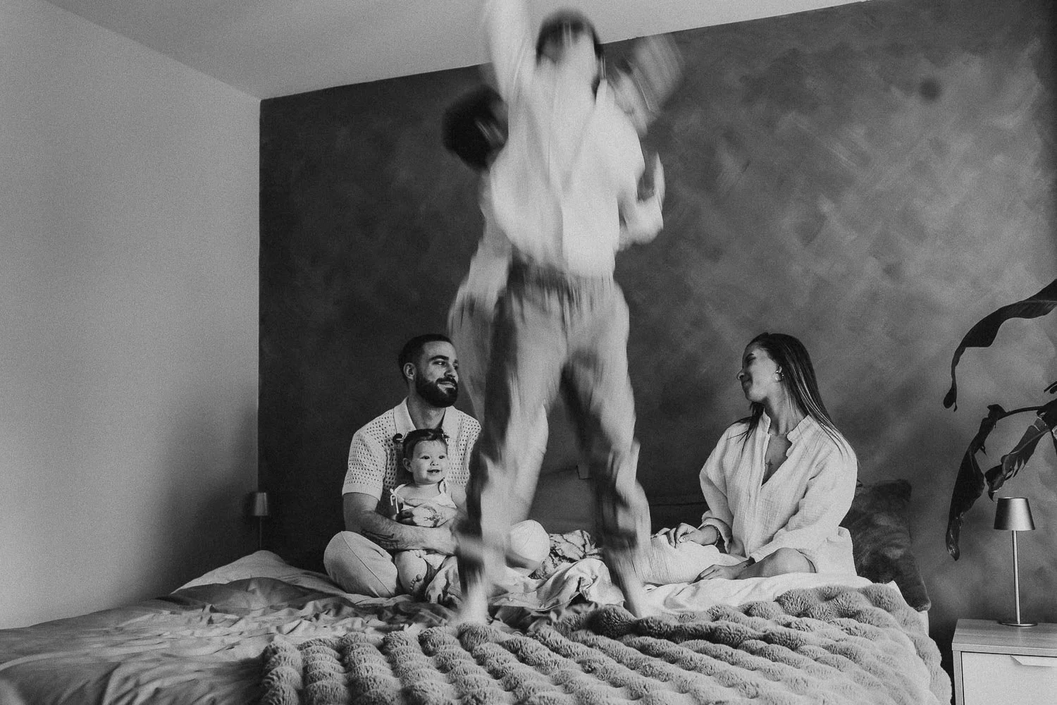 Family of five playing in bed. Mom and dad calmly looking at each other, 2 boys jumping on the bed, captured during an in-home family photoshoot in Montreal