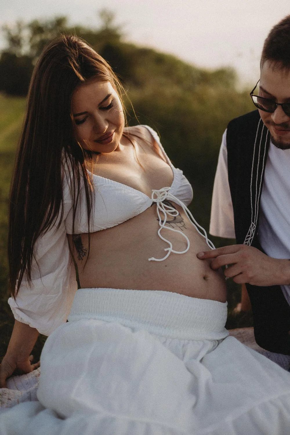 A pregnant woman seating on the floor while her partner is gently touching her belly, captured during an outdoor maternity photoshoot in Boucherville Island, Montreal