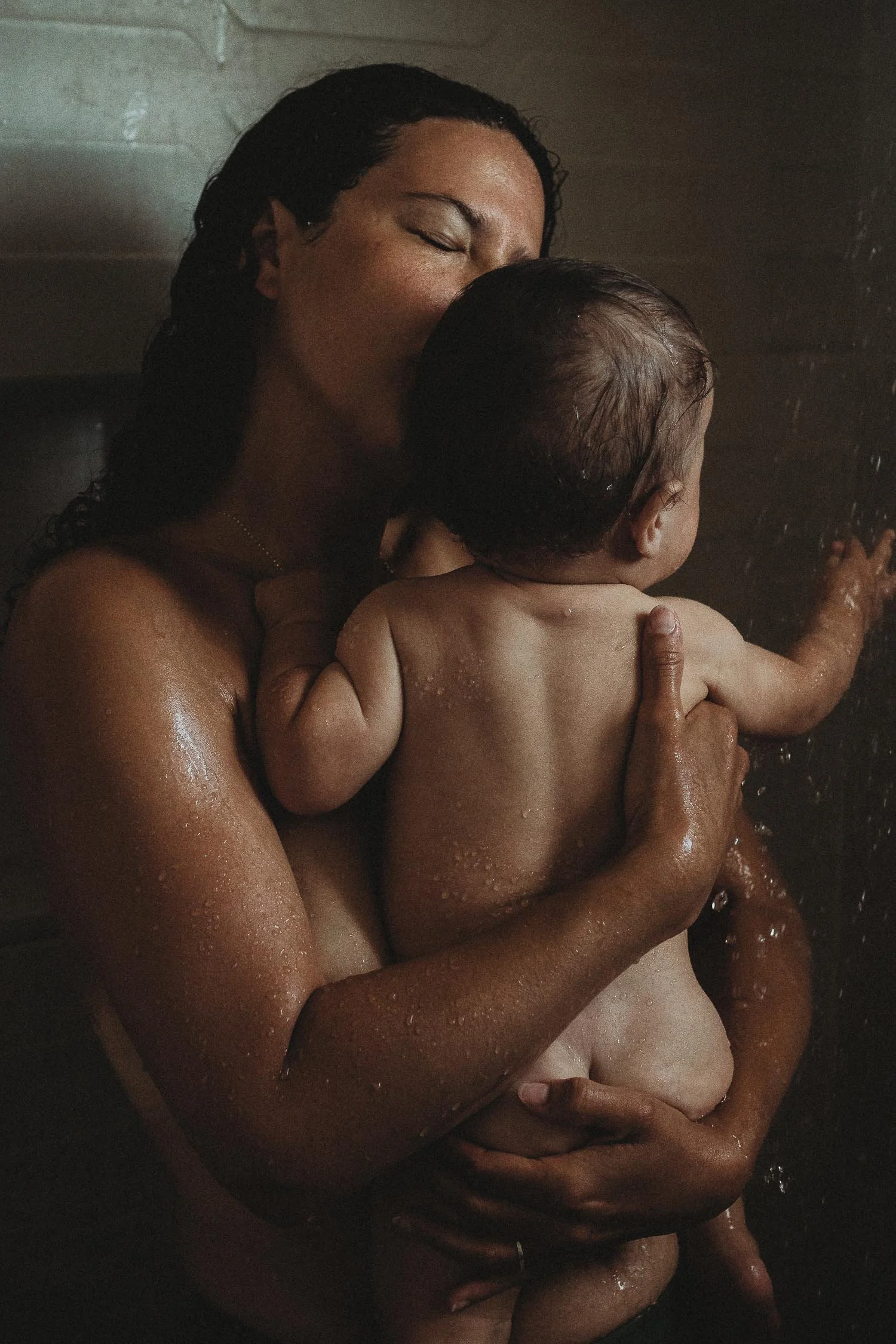 A woman holding a baby in a shower, both with wet hair and water droplets on their skin captured during an intimate Motherhood Photo Session.