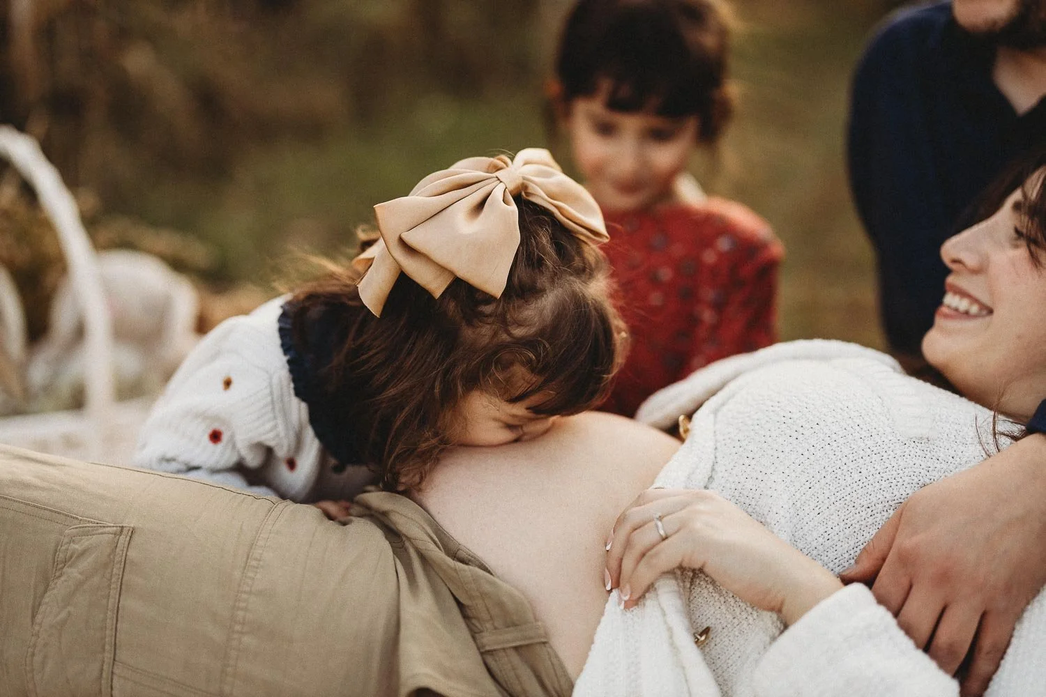A woman is lying on her back, pregnant, smiling as a young girl kisses her belly. Two children and an adult are also present, gathered outdoors on a grassy area captured at a Maternity photoshoot in Boucherville Islands, near Montreal
