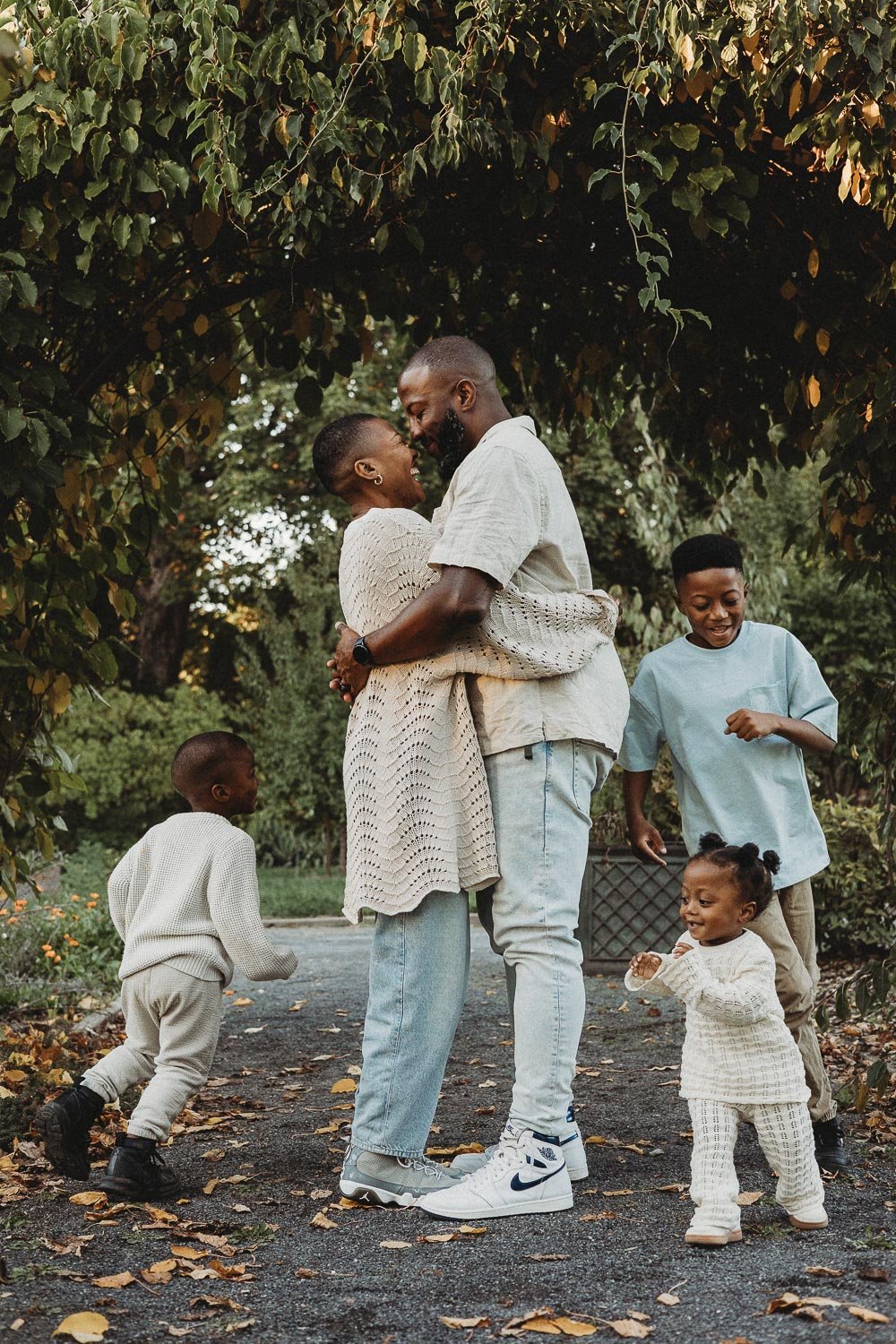 A mom and dad hugging and laughing while their 3 young kids are running around them, they are surrounded by nature in an outdoor family photoshoot in the Montreal Botanical Garden
