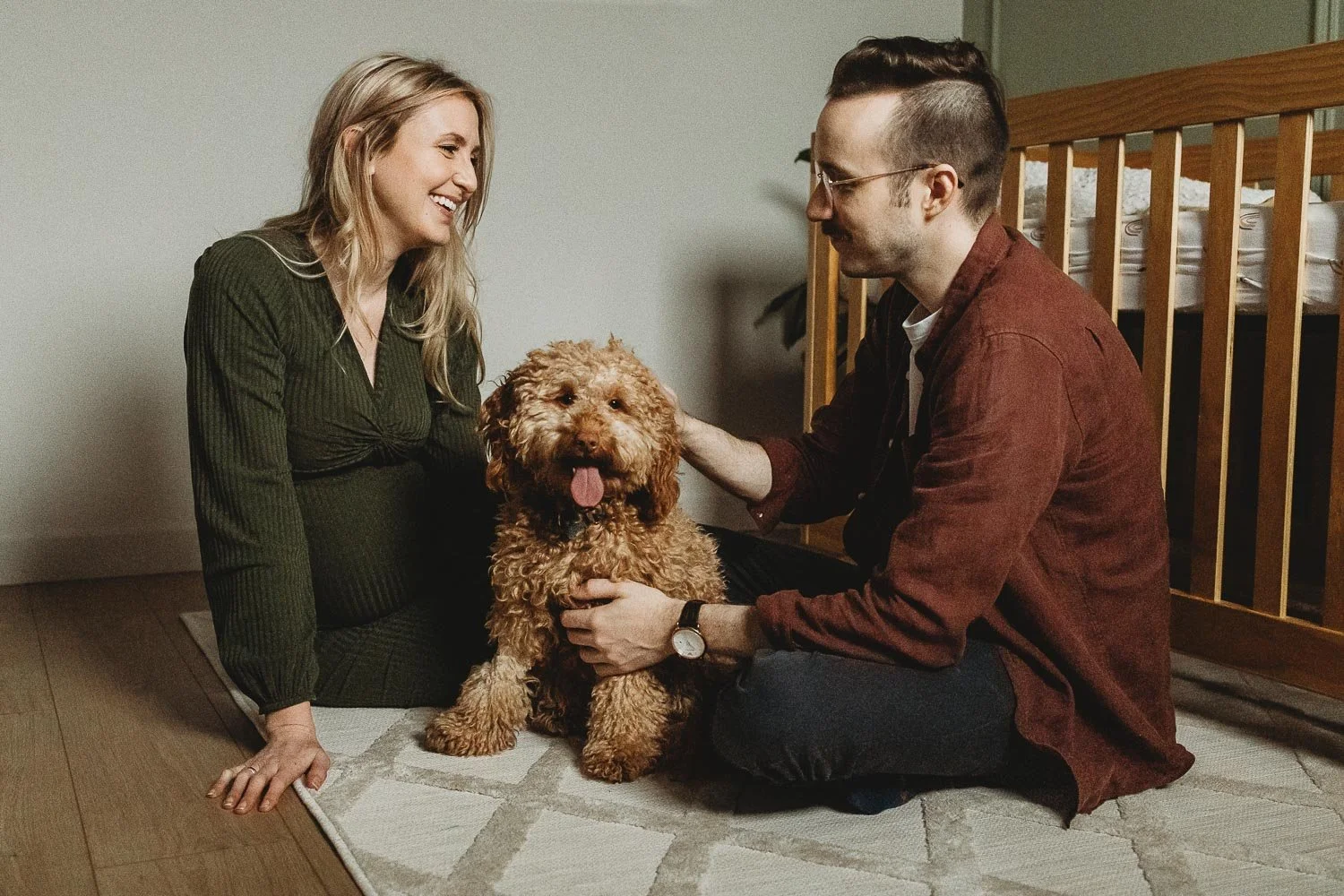 A woman and a man sitting on the floor with a curly-haired dog between them, in a room with a crib in the background. The woman is smiling and the man is petting the dog.